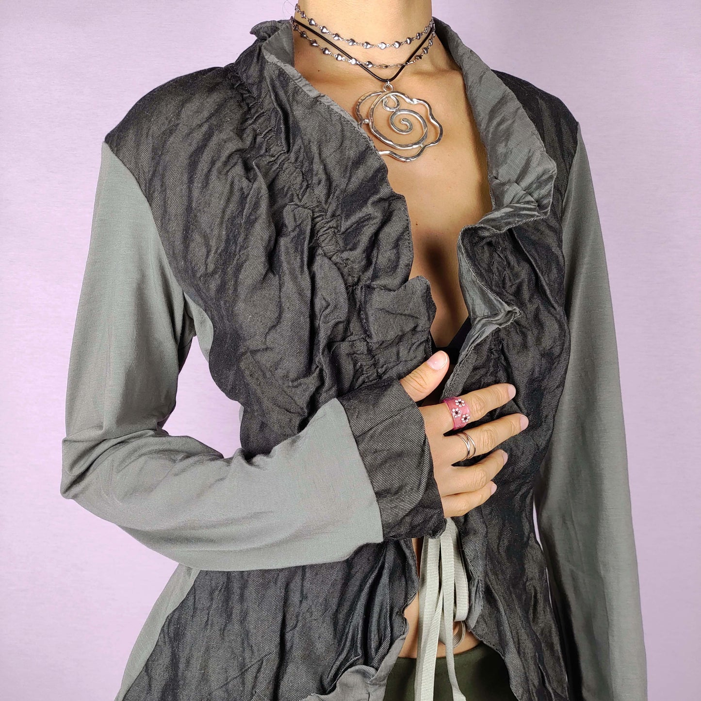 The Y2K Ruffle Tie Front Top is a vintage 2000s khaki-green grayish elegant and formal bolero jacket featuring a unique crinkled texture, ruffled details along the front and an adjustable tie closure. Perfect for both office wear and evening events.