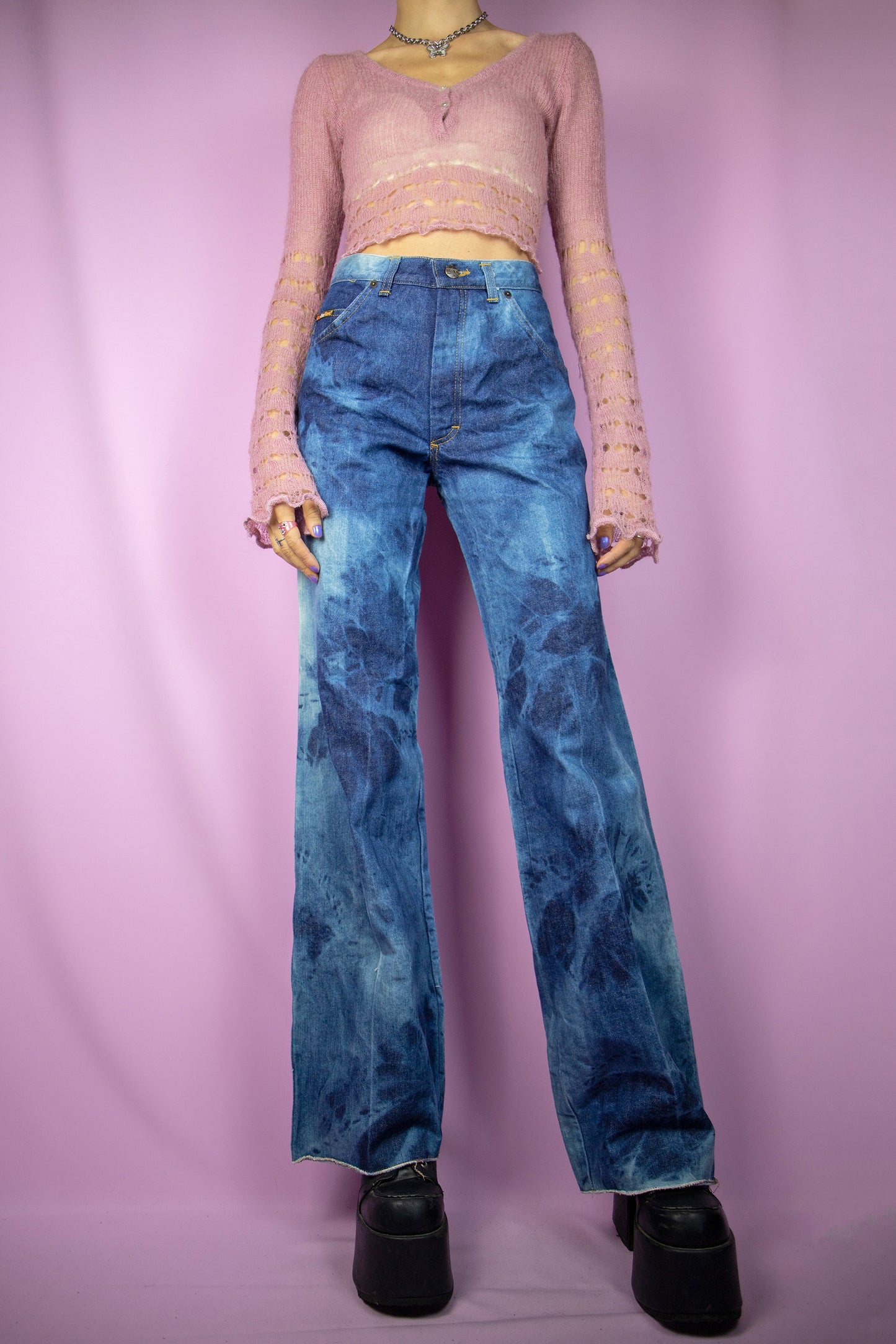 The Vintage 90s Tie Dye Wide Jeans feature a high-waisted design with a relaxed, wide-leg silhouette. These jeans showcase a bold tie-dye wash in varying shades of blue and are finished with a zipper closure, pockets, and a raw hem. Perfect for festivals and casual wear.