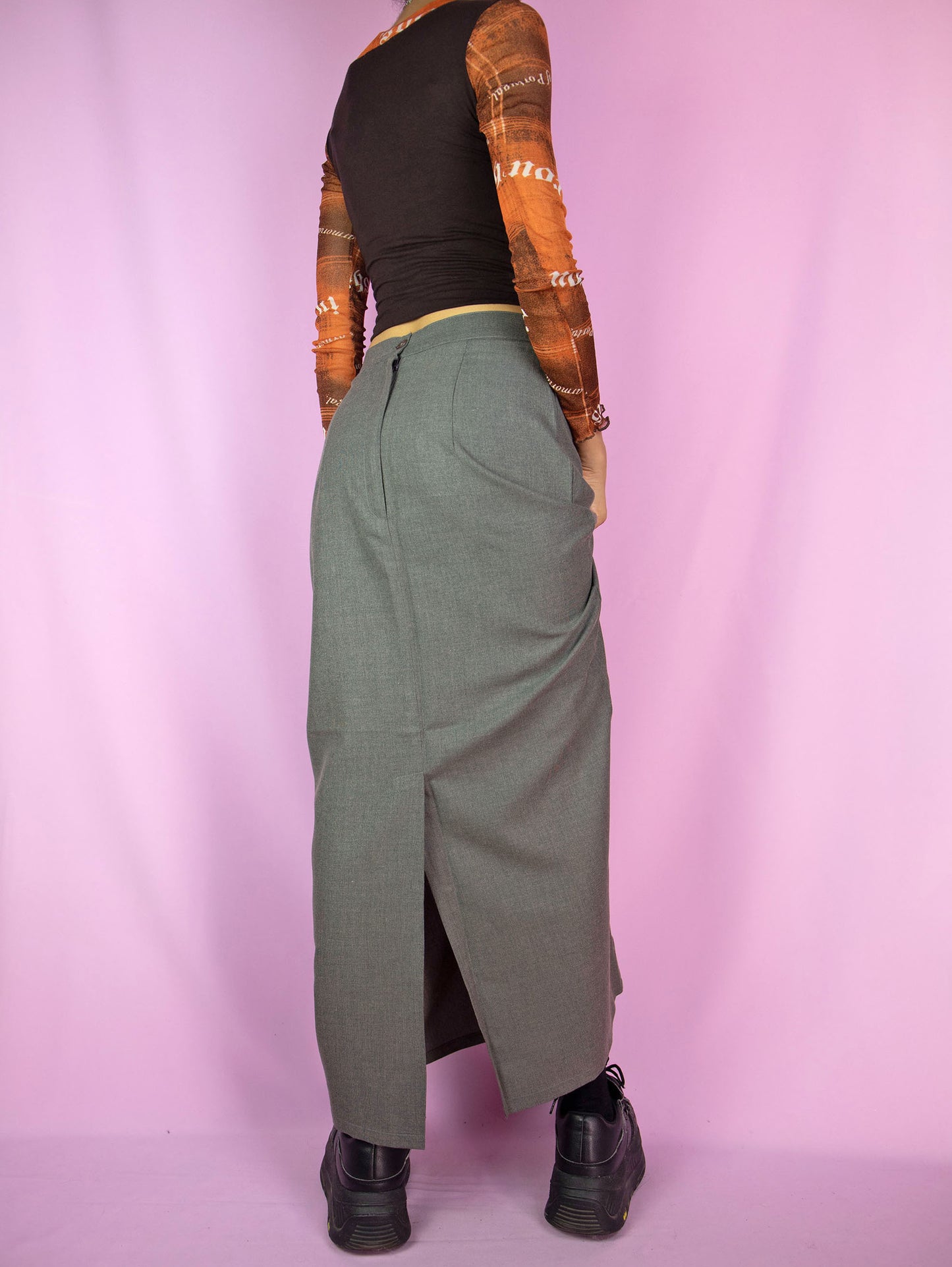 The Vintage 90s Gray Maxi Skirt features a mid-rise, A-line design with a tie-front detail, a front pocket, a back zipper closure, and a back slit. This longline skirt has a sleek, minimalist look.