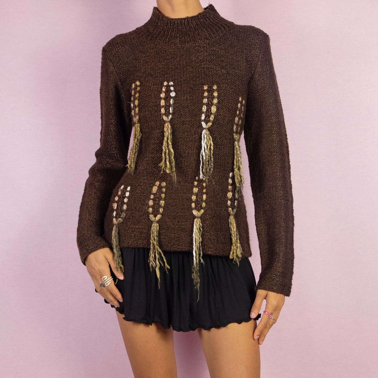 The Y2K Brown Turtleneck Sweater is a unique vintage 2000s warm pullover crafted from a cozy wool blend. It features a high turtleneck and standout embellishments with tassel details running vertically along the front. Perfect for any fall or winter wardrobe.
