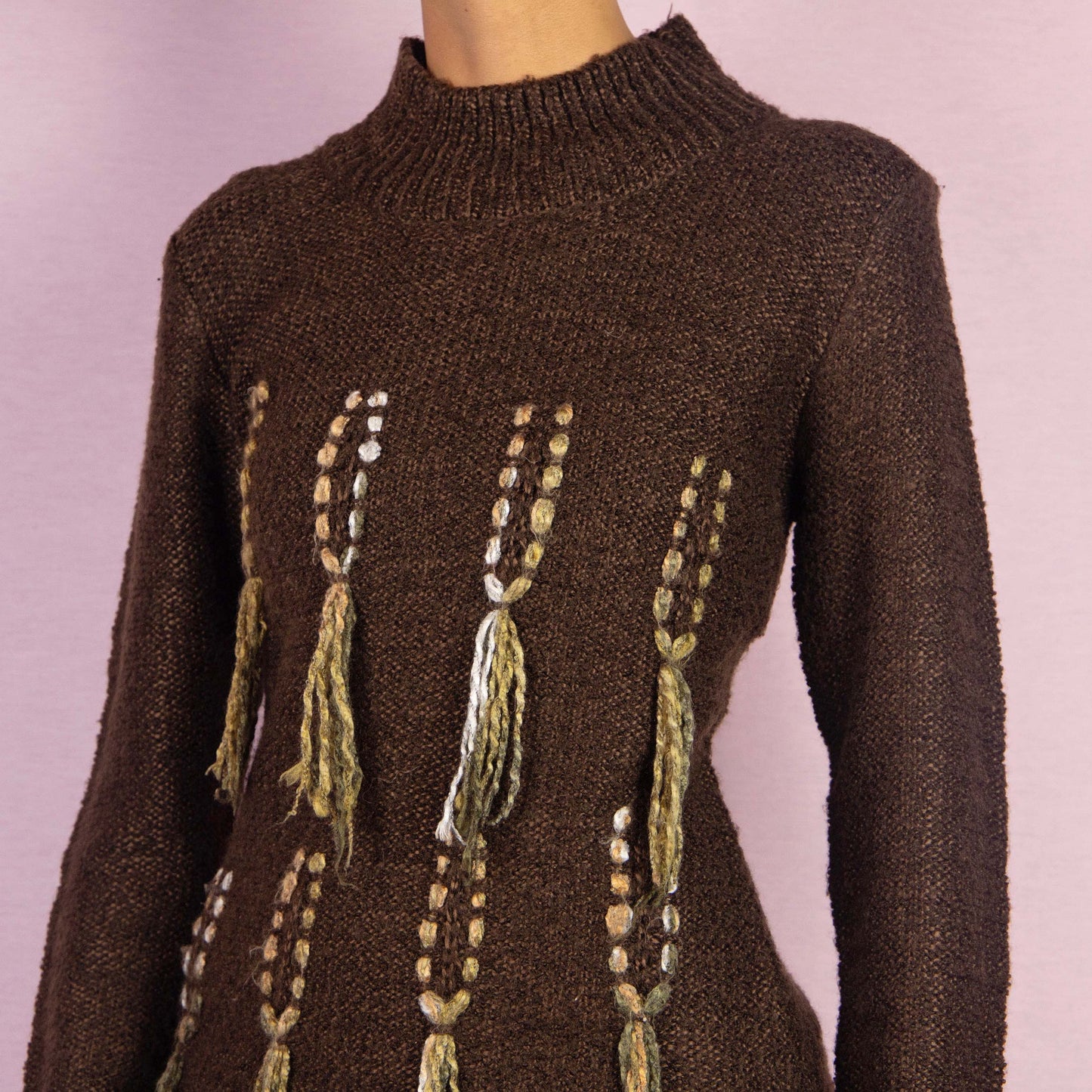 The Y2K Brown Turtleneck Sweater is a unique vintage 2000s warm pullover crafted from a cozy wool blend. It features a high turtleneck and standout embellishments with tassel details running vertically along the front. Perfect for any fall or winter wardrobe.