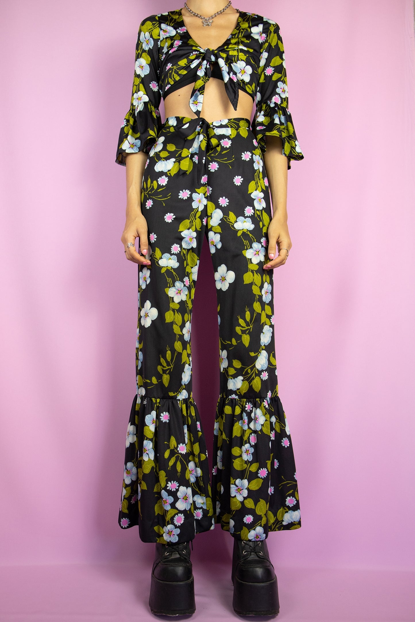 The Vintage 80s Floral Ruffle Pants Set is a statement two-piece featuring a floral print on a sleek black base. The cropped tie-front top has ruffle-trimmed bell sleeves. The matching high-waisted flared pants include a tie front zipper closure and ruffle details at the hem. Perfect for summer events, or a night out.