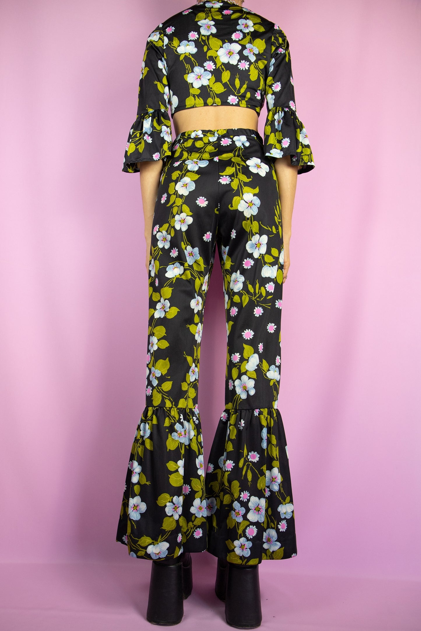 The Vintage 80s Floral Ruffle Pants Set is a statement two-piece featuring a floral print on a sleek black base. The cropped tie-front top has ruffle-trimmed bell sleeves. The matching high-waisted flared pants include a tie front zipper closure and ruffle details at the hem. Perfect for summer events, or a night out.