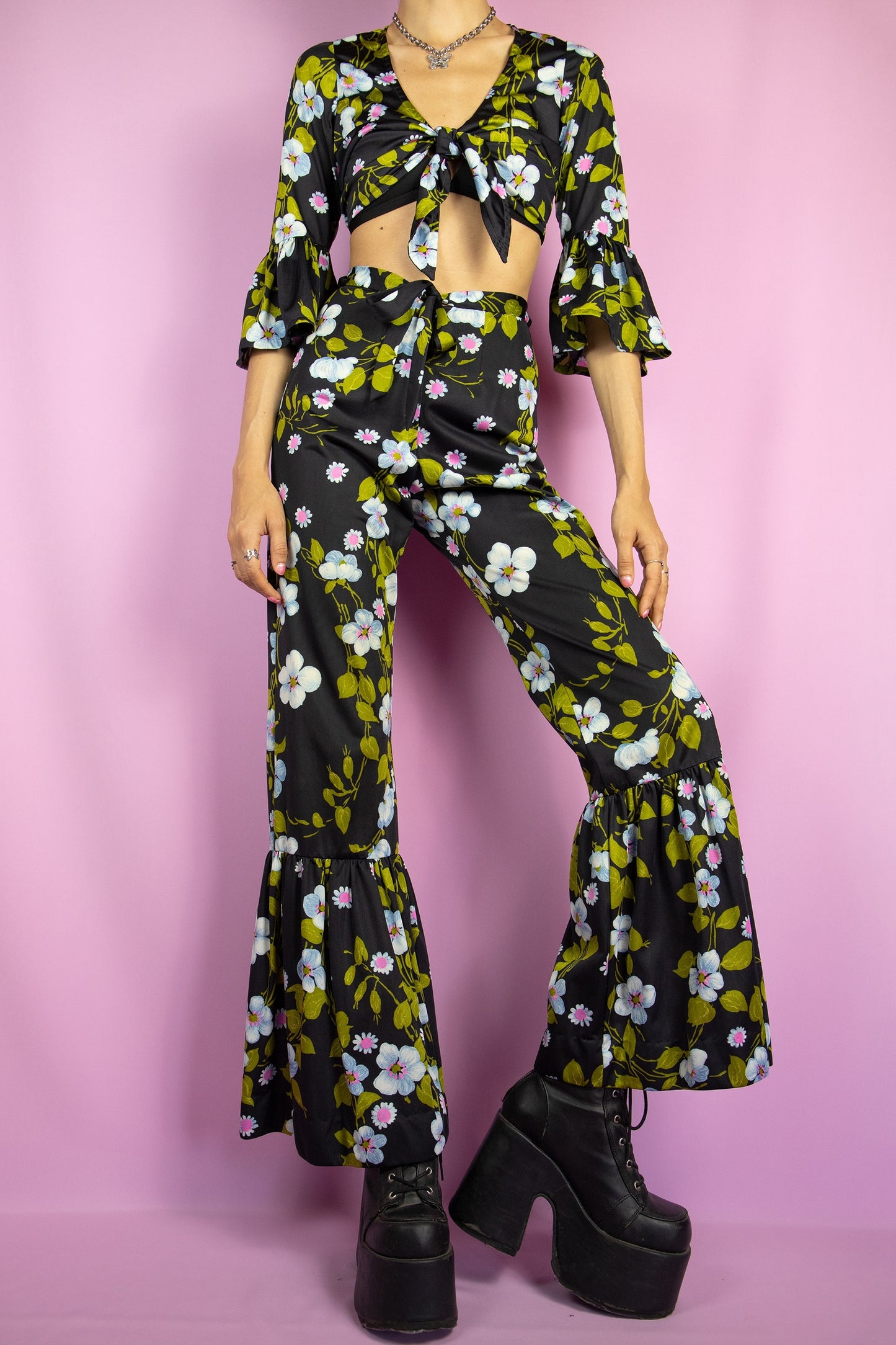 The Vintage 80s Floral Ruffle Pants Set is a statement two-piece featuring a floral print on a sleek black base. The cropped tie-front top has ruffle-trimmed bell sleeves. The matching high-waisted flared pants include a tie front zipper closure and ruffle details at the hem. Perfect for summer events, or a night out.