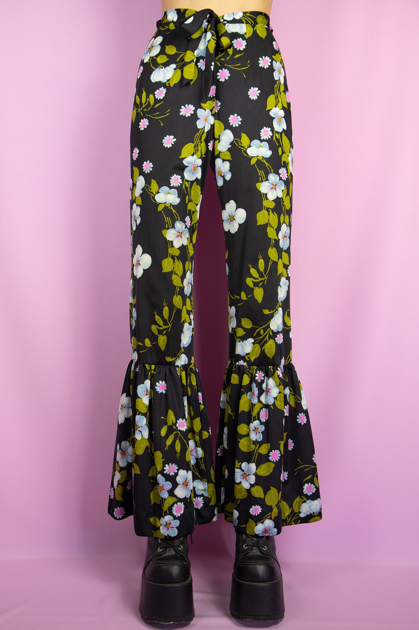 The Vintage 80s Floral Ruffle Pants Set is a statement two-piece featuring a floral print on a sleek black base. The cropped tie-front top has ruffle-trimmed bell sleeves. The matching high-waisted flared pants include a tie front zipper closure and ruffle details at the hem. Perfect for summer events, or a night out.