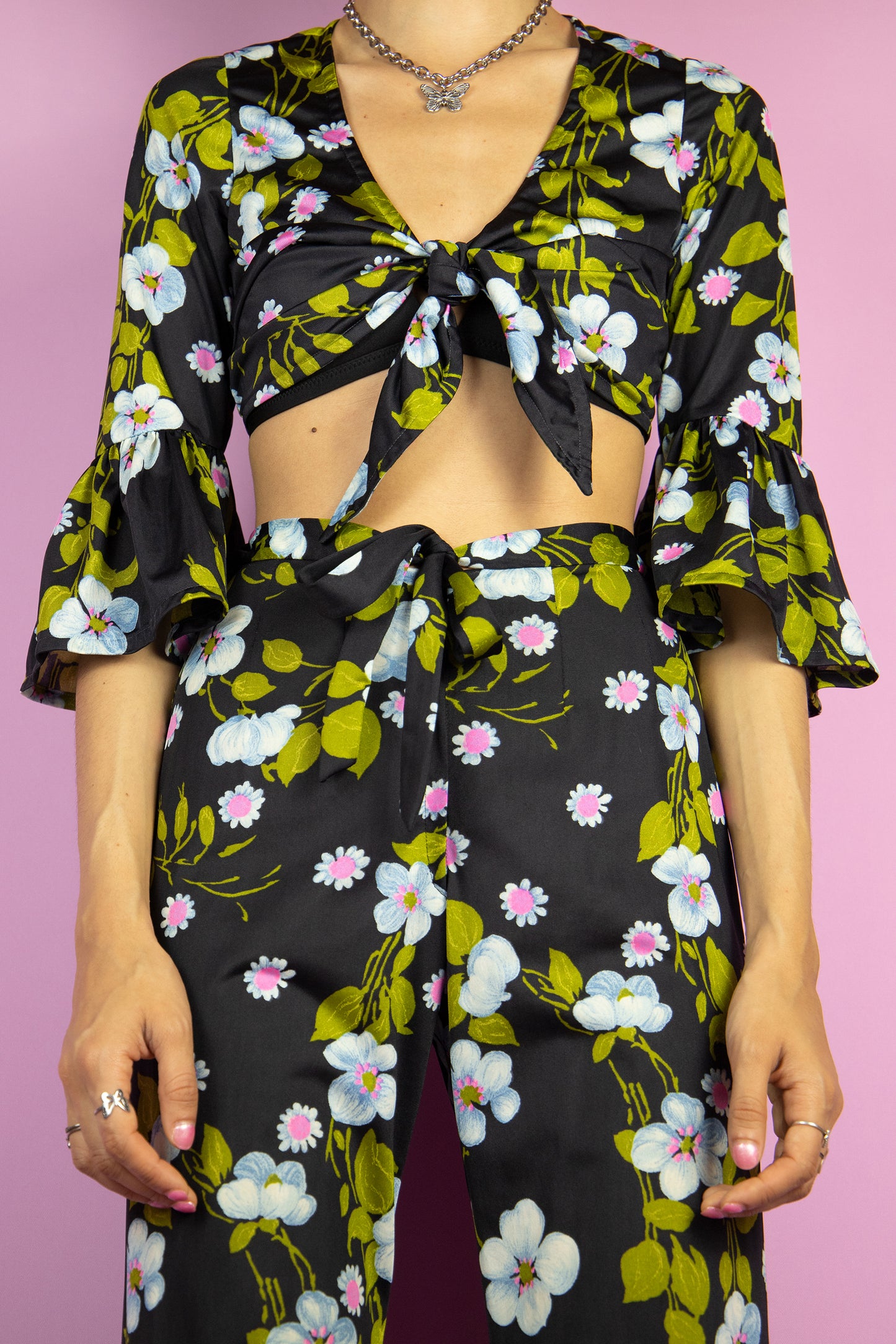 The Vintage 80s Floral Ruffle Pants Set is a statement two-piece featuring a floral print on a sleek black base. The cropped tie-front top has ruffle-trimmed bell sleeves. The matching high-waisted flared pants include a tie front zipper closure and ruffle details at the hem. Perfect for summer events, or a night out.