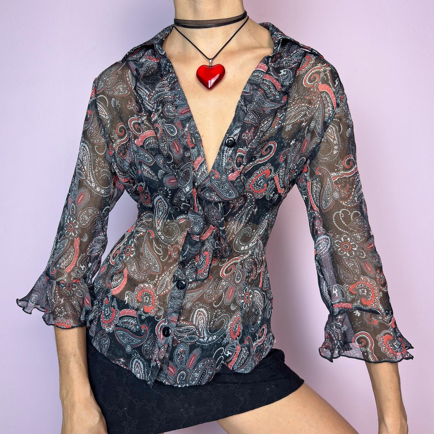 The Vintage 90s Black Ruffle Sheer Blouse features a semi-sheer fabric with an allover paisley print in red, grey, and white tones. This fitted silhouette includes three-quarter bell sleeves with flared ruffle cuffs, a romantic ruffled collar, and a front button-up closure. The lightweight fabric makes it perfect for summer layering or wearing on its own for a sleek, sexy vibe. Great vintage condition. adult world shop