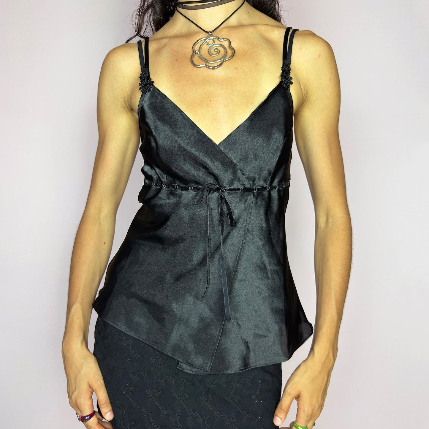 This vintage 90s black satin-style cami top features a wrap front silhouette with a subtle tie at the waist, adding gentle structure to its soft drape. Finished with delicate floral appliqués and adjustable double spaghetti straps, it captures a mood of refined sensuality. Perfect for cocktail nights, evening events, or romantic layering. Lightweight and fluid, it brings feminine elegance with a hint of 90s glamour. Excellent vintage condition.