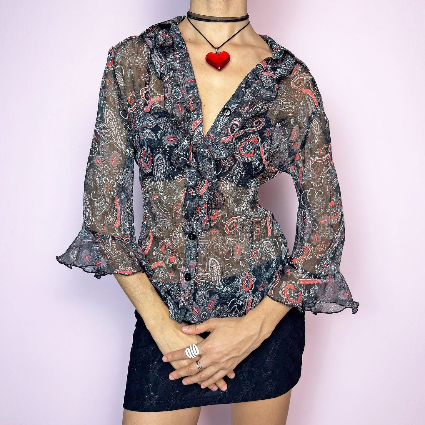 The Vintage 90s Black Ruffle Sheer Blouse features a semi-sheer fabric with an allover paisley print in red, grey, and white tones. This fitted silhouette includes three-quarter bell sleeves with flared ruffle cuffs, a romantic ruffled collar, and a front button-up closure. The lightweight fabric makes it perfect for summer layering or wearing on its own for a sleek, sexy vibe. Great vintage condition.