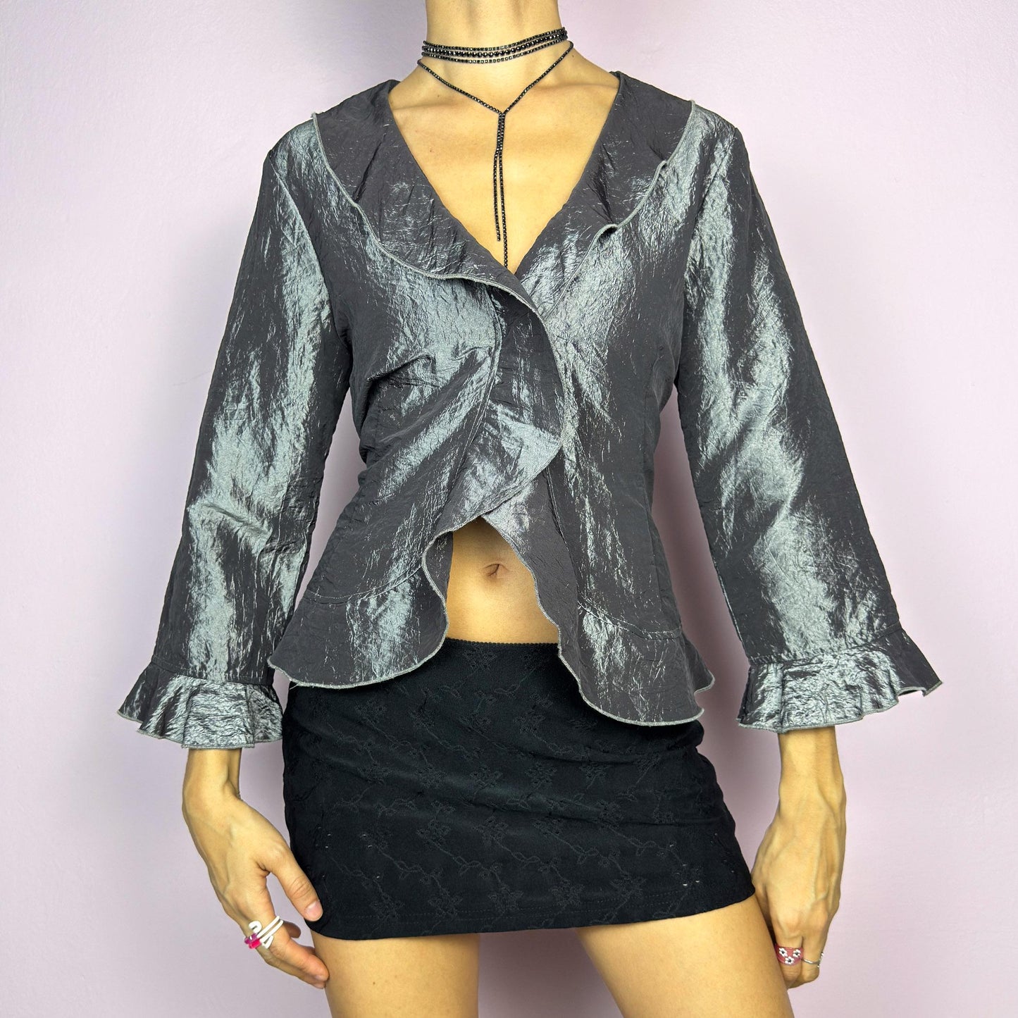 The Vintage 90s Gray Iridescent Ruffle Blouse is a sophisticated and elegant piece with a shimmering iridescent finish in a metallic gray tone. It features a crinkled texture, a cascading ruffle trims along the hemline. adult world shop
