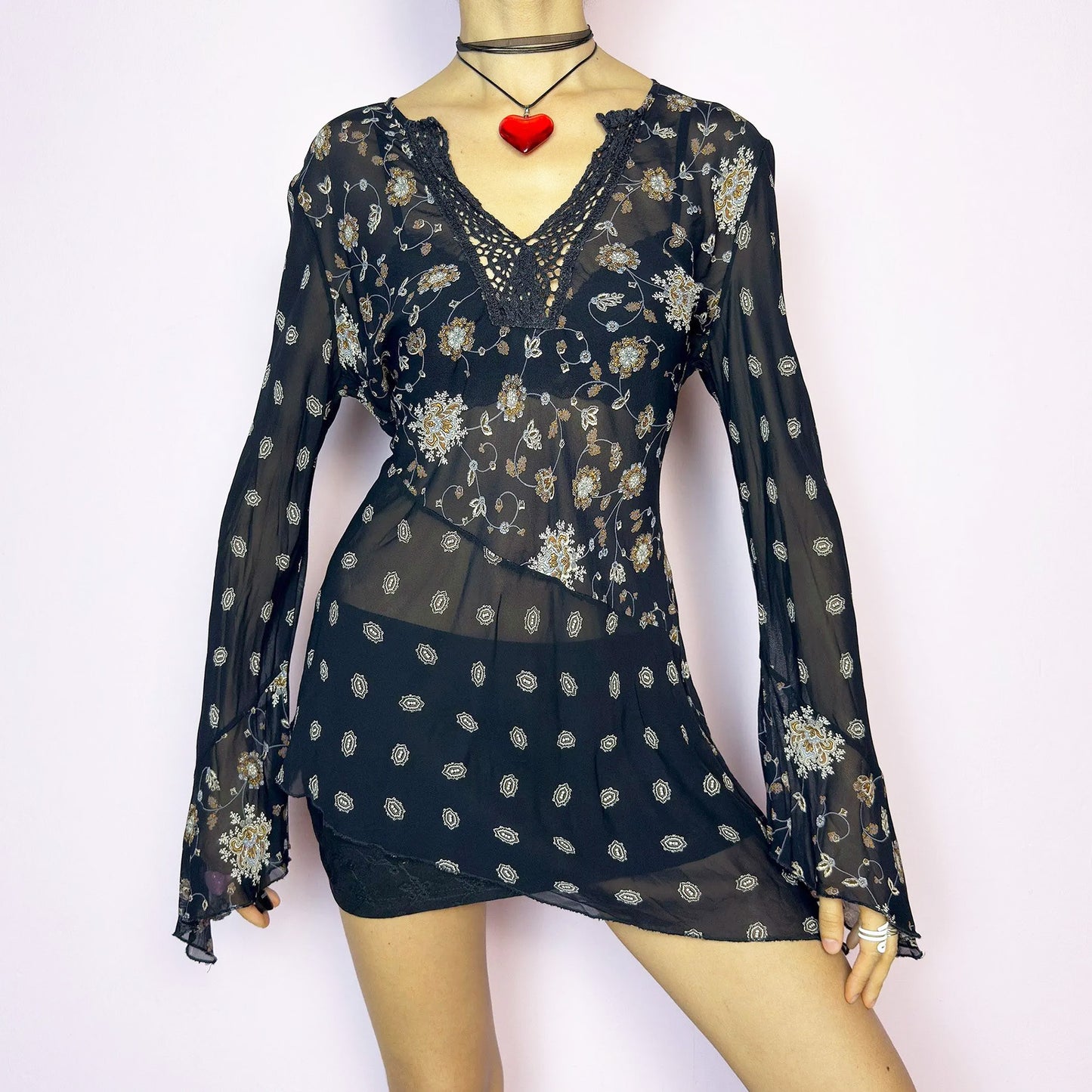 The Y2K Asymmetric Bell Sleeve Top is a vintage 2000s piece featuring a sheer black floral print with an asymmetric hem and dramatic bell sleeves for a flowy silhouette. The deep V-neckline is accented with crochet lace trim for a romantic touch. Lightweight and breathable, it’s perfect for summer layering or festival styling. Includes long sleeves with a bell flare for an elegant witchy vibe. Great vintage condition.