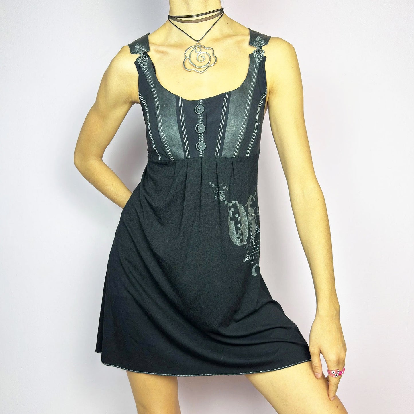 The Y2K Black Buckle Strap Mini Dress is a vintage 2000s sleeveless piece with a mix of edgy and casual aesthetics. It features decorative button details, contrast stitching, and graphic prints. The standout buckle strap details add an industrial and cyber vibe. Made from a stretchy fabric, it offers a comfortable fit, perfect for casual streetwear or alternative fashion looks.