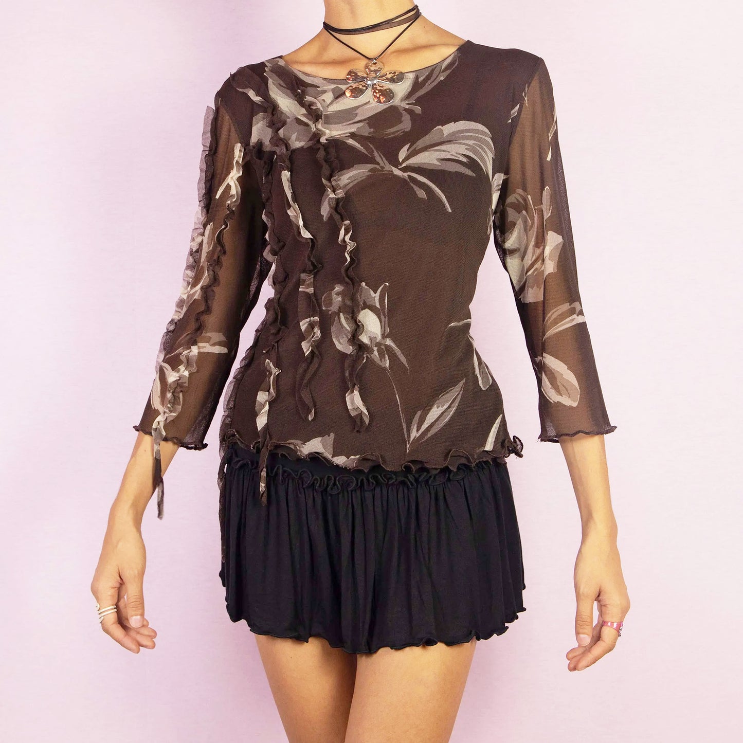 The Y2K Brown Frill Mesh Top is a vintage 2000s blouse featuring a semi-sheer fabric with a floral print in earthy brown tones. The top is detailed with whimsical ruffle accents along the front and sleeves, an asymmetric design, and three-quarter sleeves. Perfect for casual or evening looks.