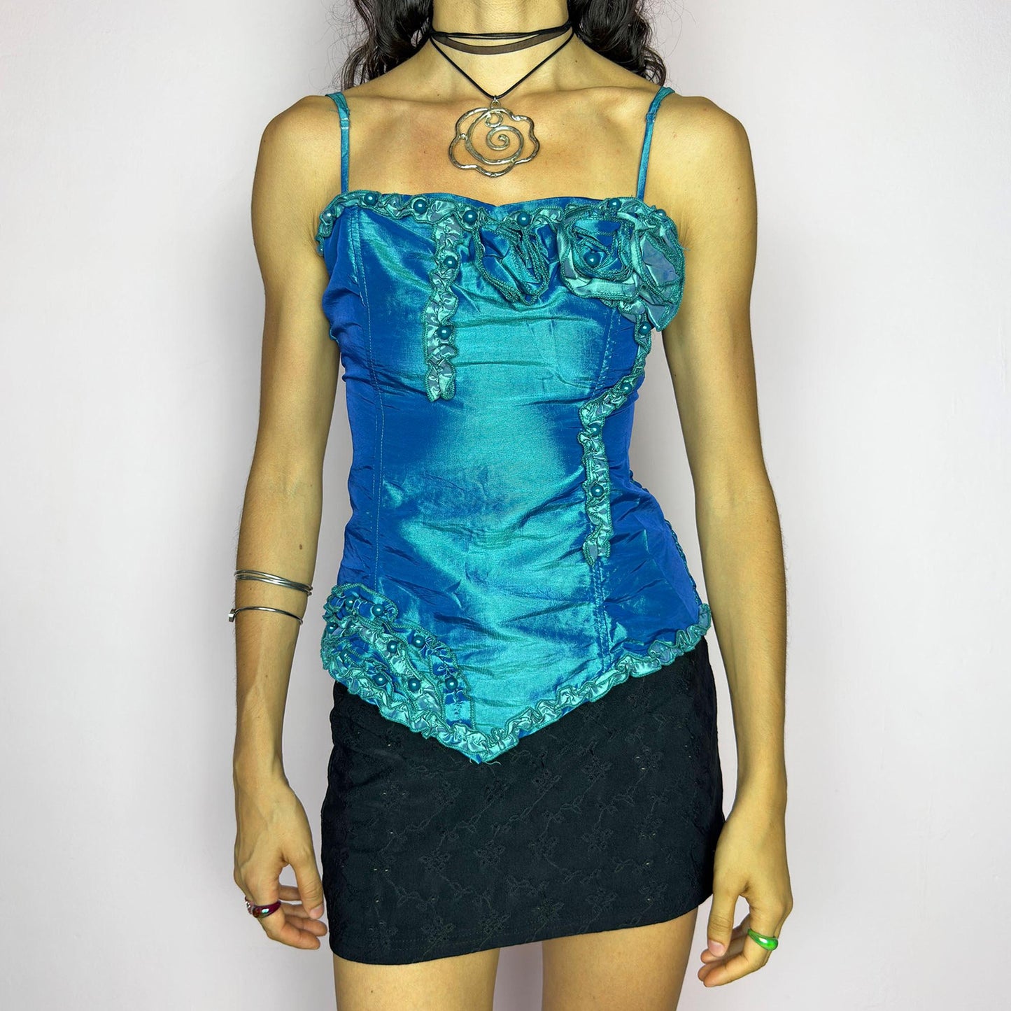 This vintage 2000s iridescent turquoise corset top features adjustable spaghetti straps, floral rosette appliqué, and beaded trim. The structured front sculpts the waist, while the ruched smocked back adds stretch and comfort. An asymmetrical hem and glossy finish elevate it for prom, weddings, or dreamy nights out. A romantic Y2K formal top that blends fairytale opulence with a bold, sculptural silhouette. Excellent vintage condition.