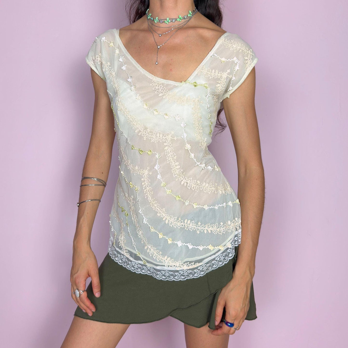 This vintage 2000s cream white mesh top features semi-sheer fabric with delicate embroidery in soft yellow, white, and pale green. The asymmetrical neckline and cap sleeves create an ethereal drape, while lace trim at the hem adds a romantic finish. This fairy-like Y2K blouse evokes lightness and whimsy—perfect for dreamy summer looks, warm days, and delicate layering. Made of polyester blend.