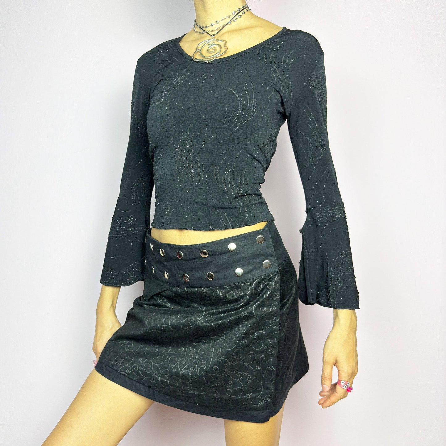 The Y2K Wrap Mini Black Skirt is a vintage 2000s piece featuring a snap-button closure on the waistband for an adjustable fit. Made from a thick fabric with a subtle floral pattern design, its wrap silhouette makes it perfect for casual outfits or festival looks.