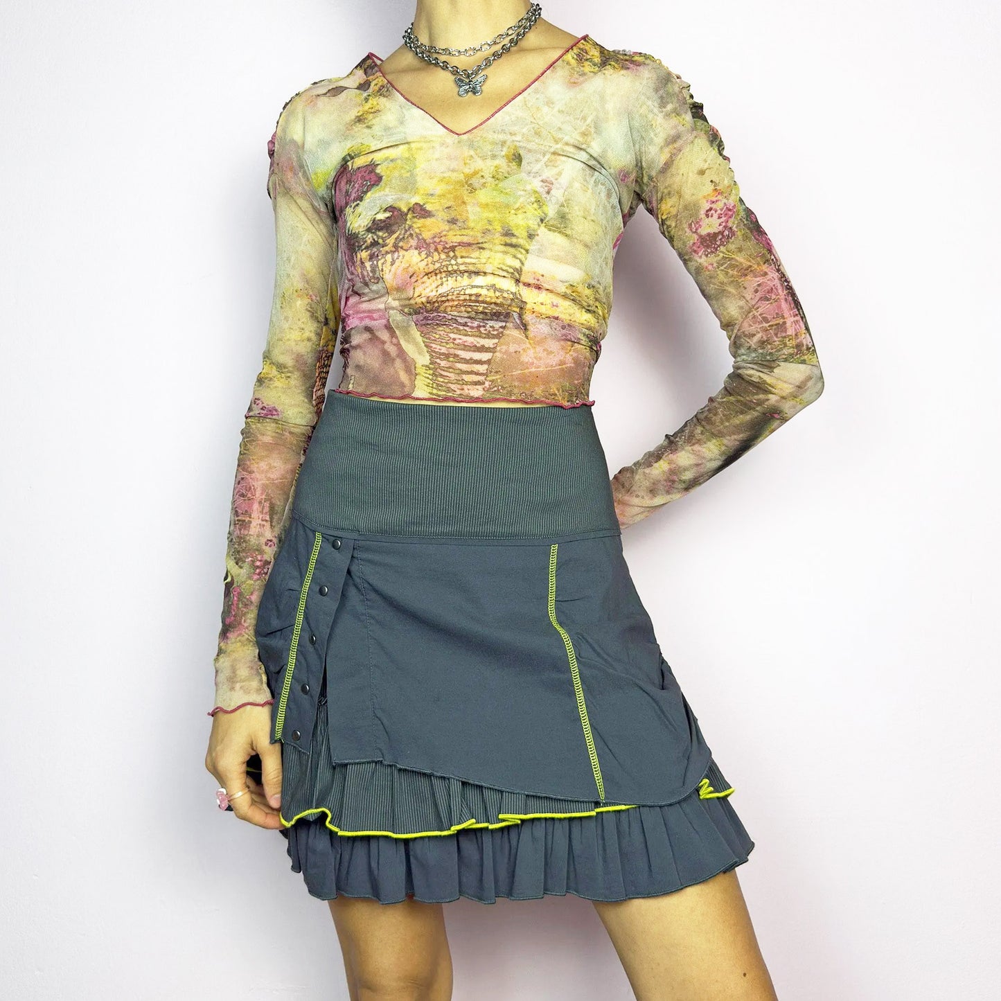 The Y2K Gray Ruffle Mini Skirt is a vintage 2000s statement piece featuring a tiered layered design with yellow contrast stitching details. Designed with a high-waist fit, it includes a side zipper closure and an adjustable ruched tie drawstring detail on the side. Perfect for cyber, techwear, or streetwear-inspired outfits.