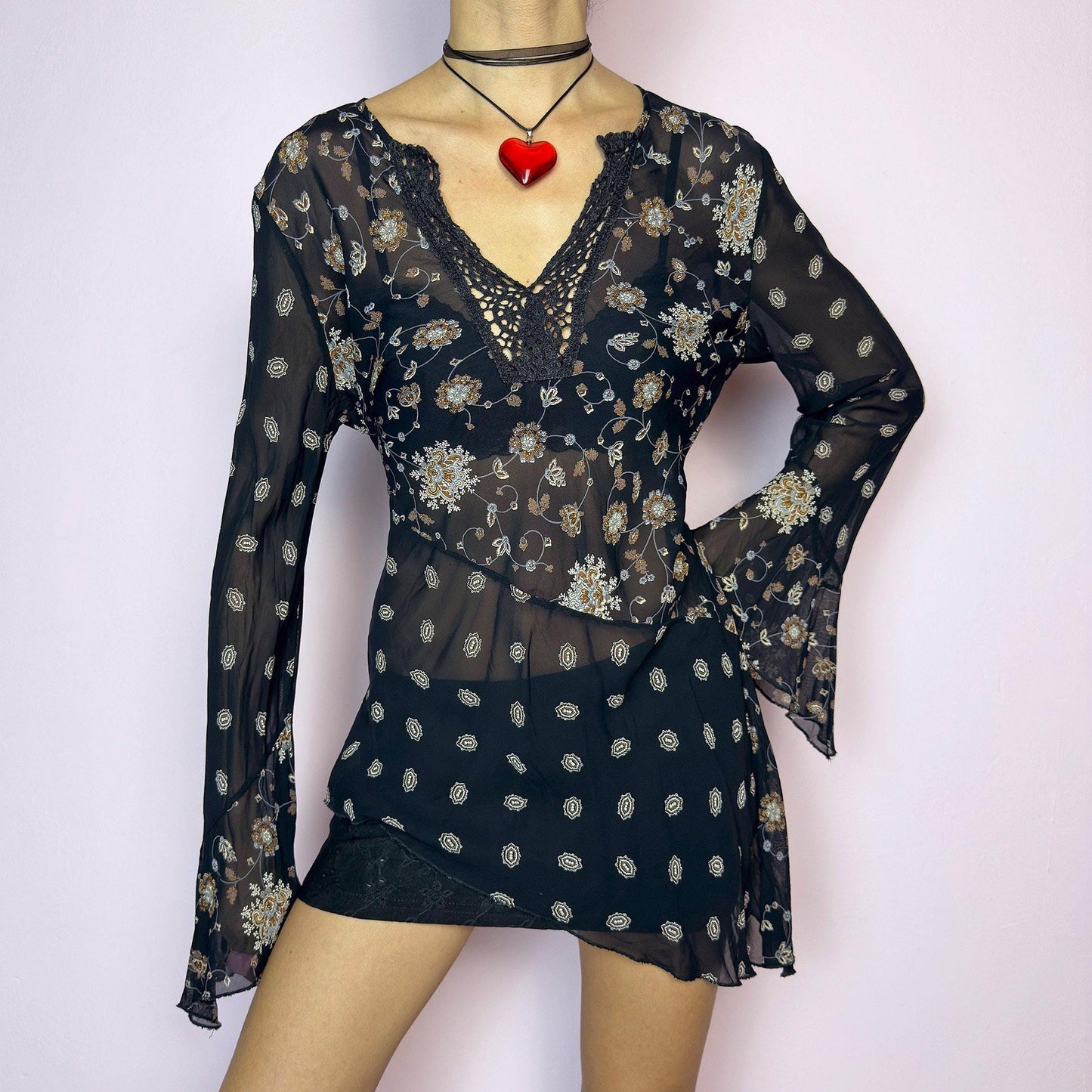 The Y2K Asymmetric Bell Sleeve Top is a vintage 2000s piece featuring a sheer black floral print with an asymmetric hem and dramatic bell sleeves for a flowy silhouette. The deep V-neckline is accented with crochet lace trim for a romantic touch. Lightweight and breathable, it’s perfect for summer layering or festival styling. Includes long sleeves with a bell flare for an elegant witchy vibe. Great vintage condition. adult world shop