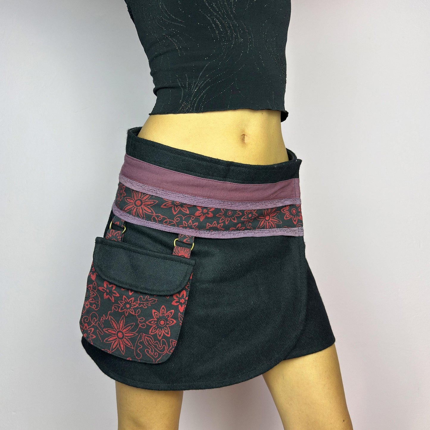 The Y2K Black Wrap Mini Skirt is a vintage 2000s piece featuring a unique wrap design with an adjustable Velcro waist for a customizable fit. Made from a thick fabric, the skirt has a contrasting waistband with floral details. adult world shop