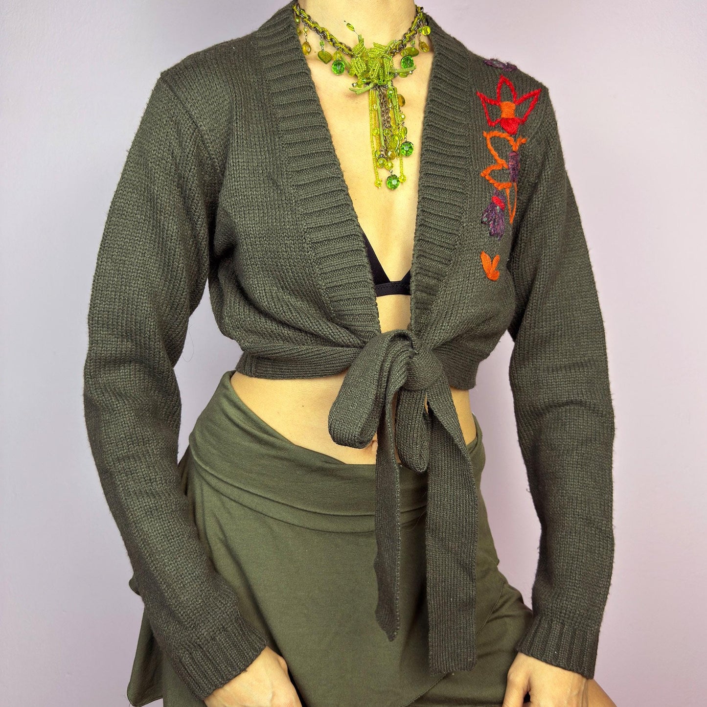 The Y2K Khaki Tie Front Cardigan is a vintage 2000s knit featuring a deep V-neckline, front tie closure, and long sleeves. Designed in a dark green hue, this cropped cardigan is accented with floral embroidery on the shoulder for a boho-chic touch. The stretchy, cozy knit makes it perfect for layering over casual or fairy grunge-inspired outfits. adult world shop