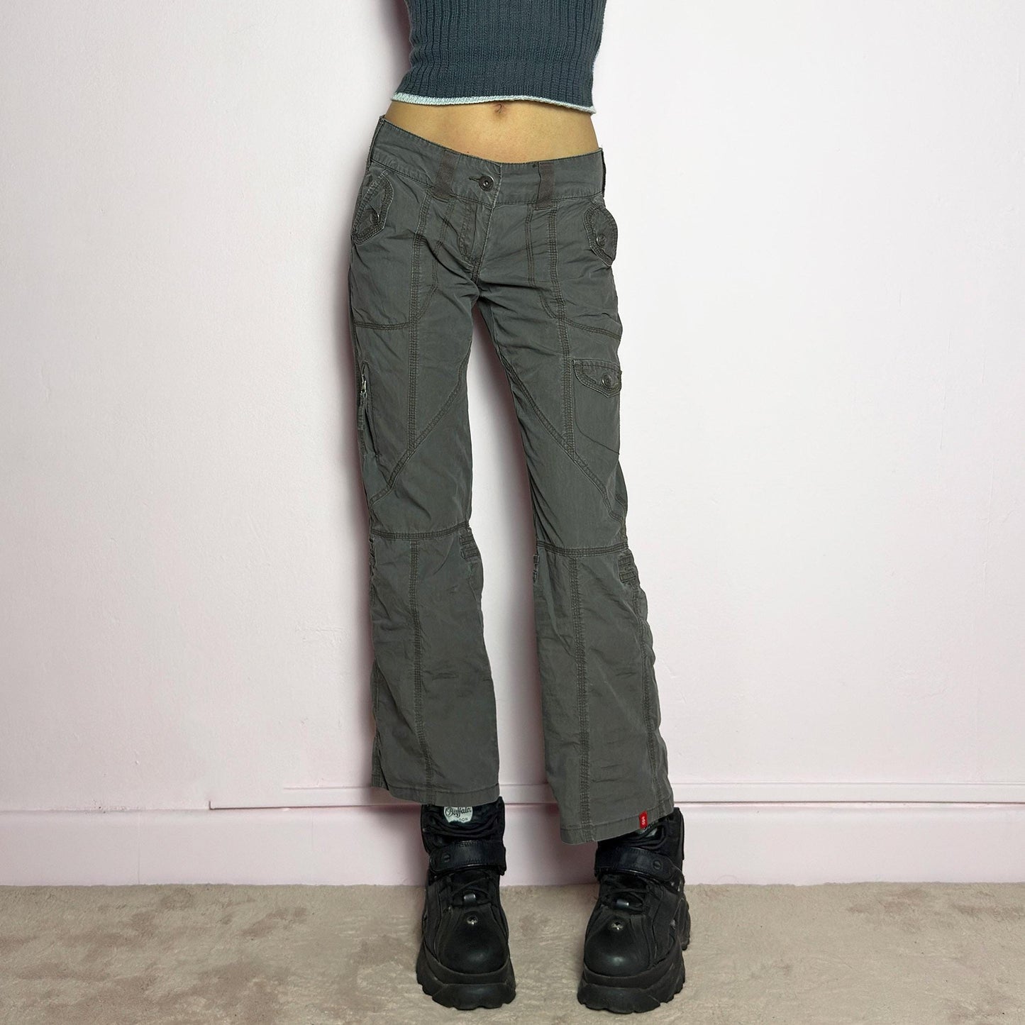 The Y2K Low-Rise Cargo Pants are a vintage 2000s piece featuring a washed gray tone with contrast stitching. Designed with a low-rise fit, button and zipper closure, and multiple cargo-style pockets, these pants have panel detailing and exposed seams that enhance their utilitarian aesthetic. The cropped straight-leg cut adds a modern touch, making them perfect for grunge and streetwear-inspired outfits. adult world shop