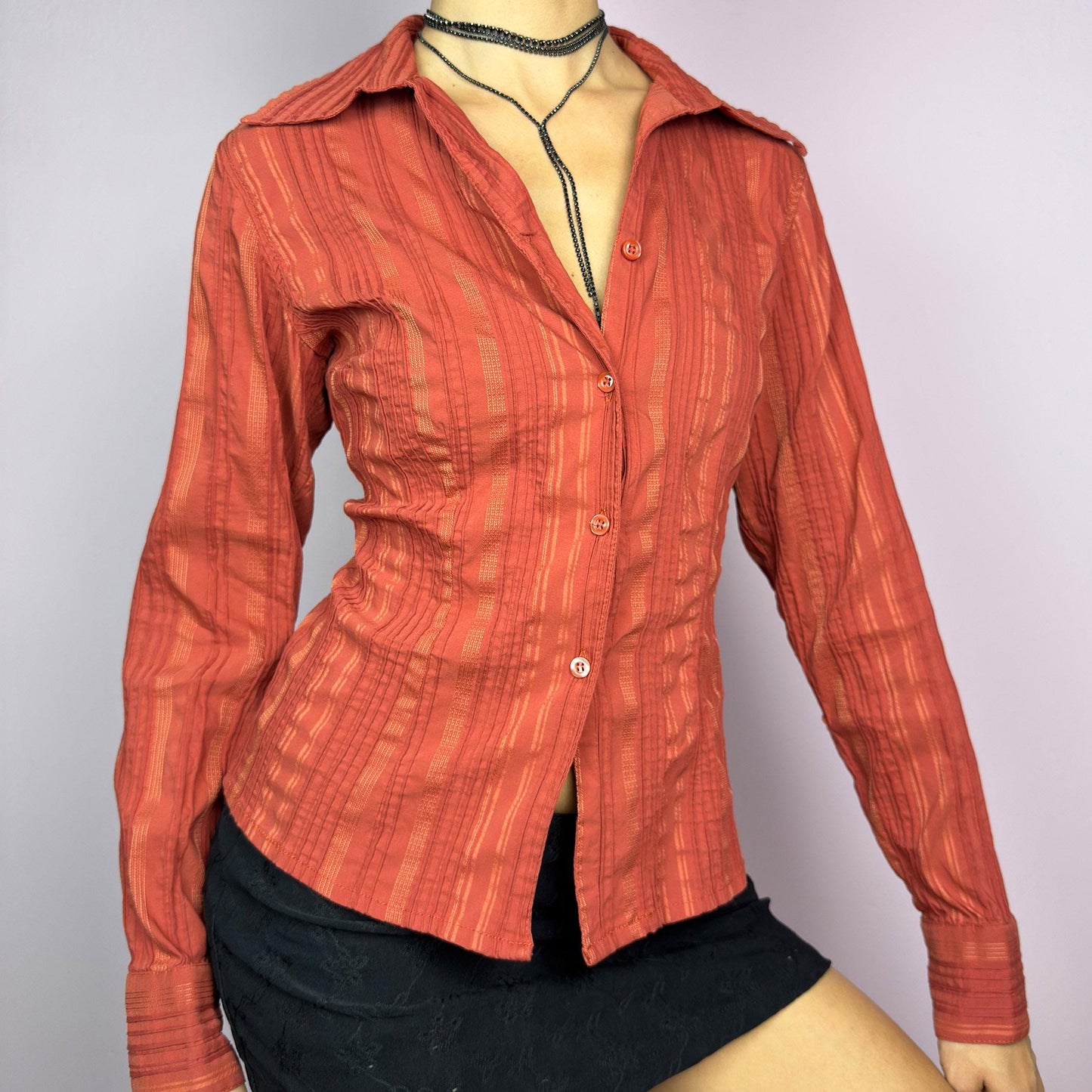 The Y2K Orange Striped Blouse is a vintage 2000s shirt featuring a warm rust-orange hue with a subtle striped pattern, a flattering tailored fit, and a hint of stretch. This long-sleeve button-up blouse is perfect for office wear. adult world shop