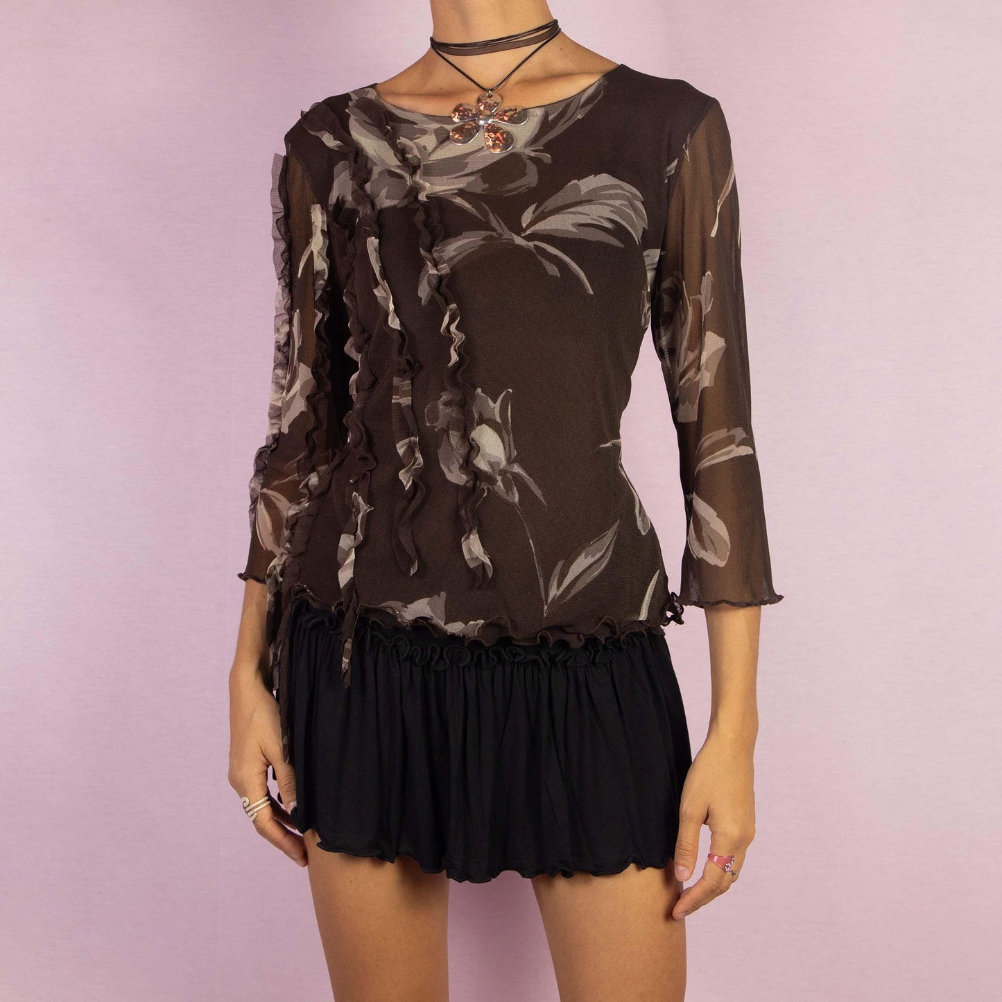 The Y2K Brown Frill Mesh Top is a vintage 2000s blouse featuring a semi-sheer fabric with a floral print in earthy brown tones. The top is detailed with whimsical ruffle accents along the front and sleeves, an asymmetric design