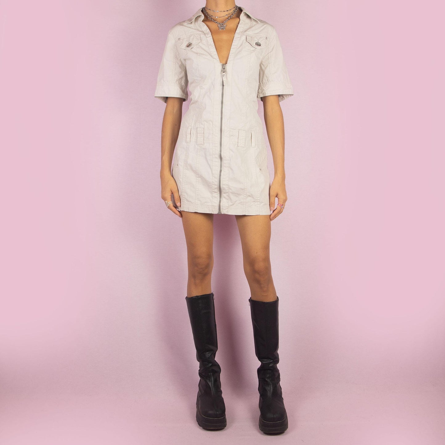 The Y2K Beige Cargo Mini Dress is a vintage 2000s piece featuring a sleek design with a front zipper closure, short sleeves, a collared neckline, and functional details like front pockets giving it a structured look. Crafted in a lightweight beige