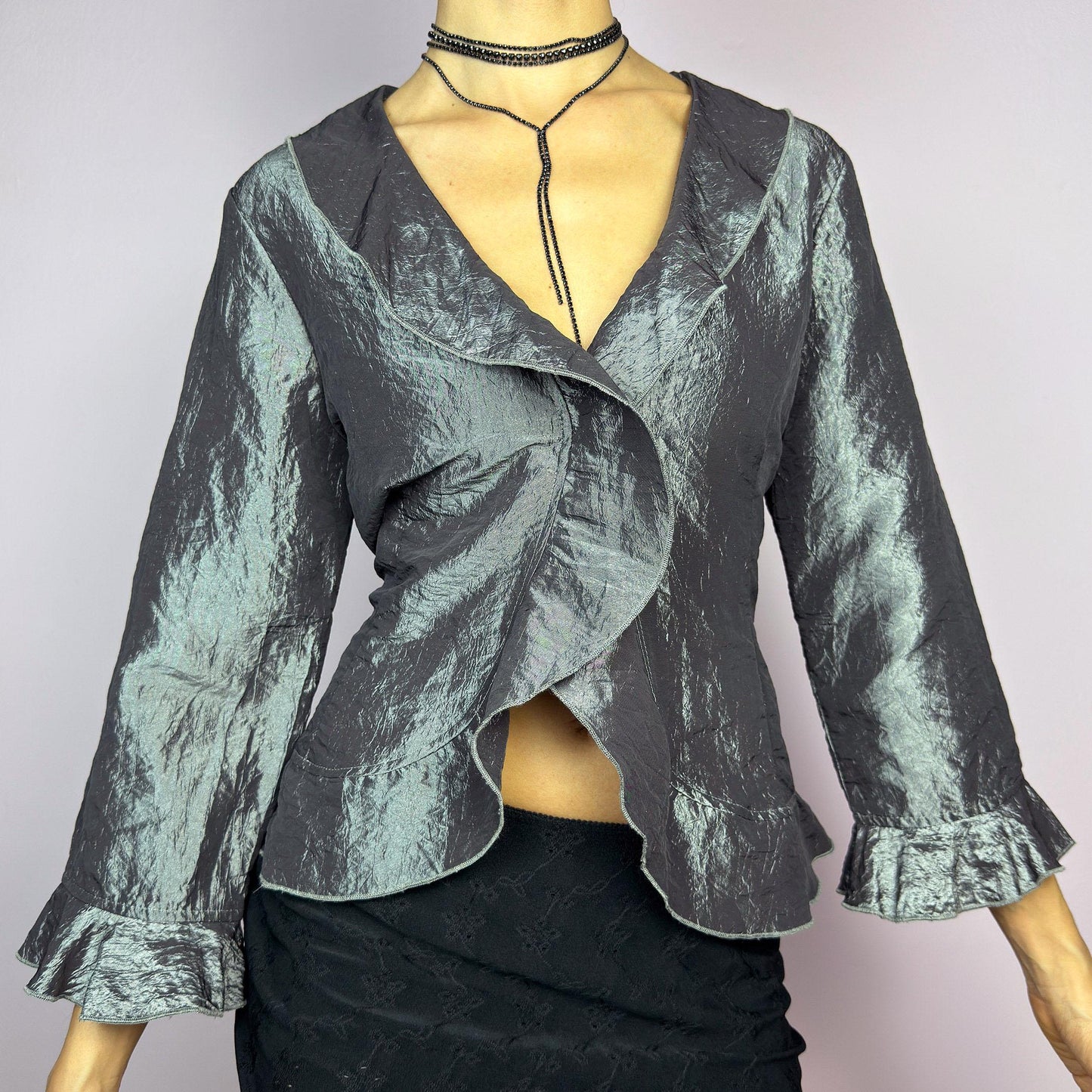 The Vintage 90s Gray Iridescent Ruffle Blouse is a sophisticated and elegant piece with a shimmering iridescent finish in a metallic gray tone. It features a crinkled texture, a cascading ruffle trims along the hemline. adult world shop