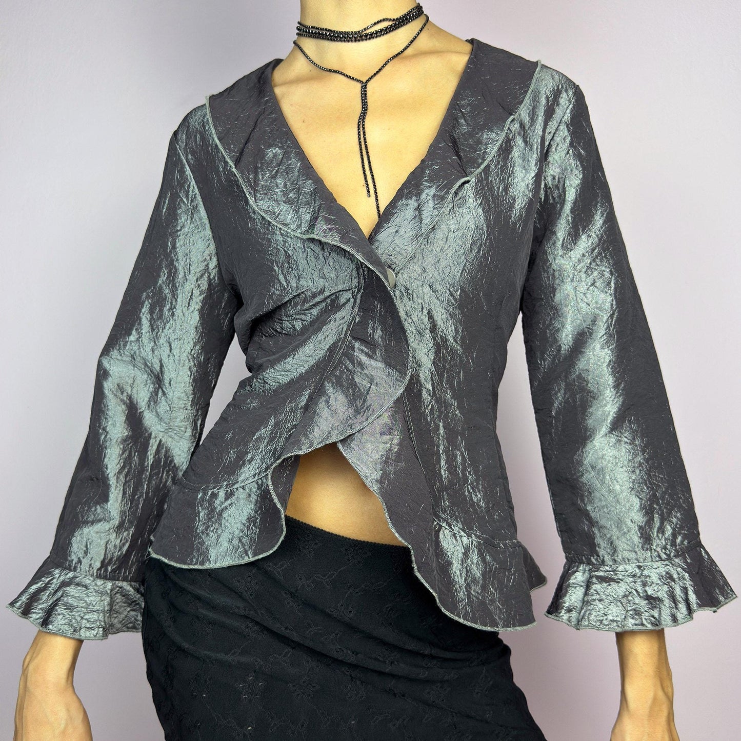 The Vintage 90s Gray Iridescent Ruffle Blouse is a sophisticated and elegant piece with a shimmering iridescent finish in a metallic gray tone. It features a crinkled texture, a cascading ruffle trims along the hemline. adult world shop