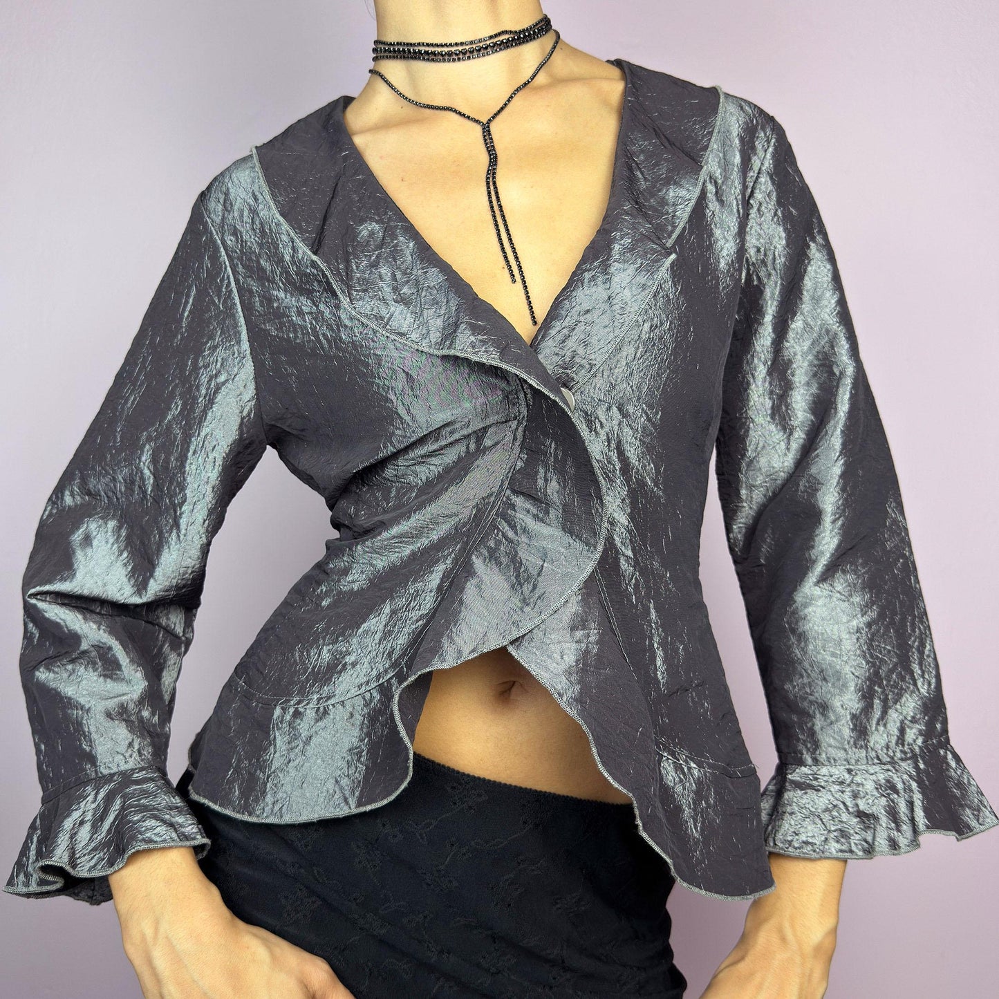 The Vintage 90s Gray Iridescent Ruffle Blouse is a sophisticated and elegant piece with a shimmering iridescent finish in a metallic gray tone. It features a crinkled texture, a cascading ruffle trims along the hemline. adult world shop