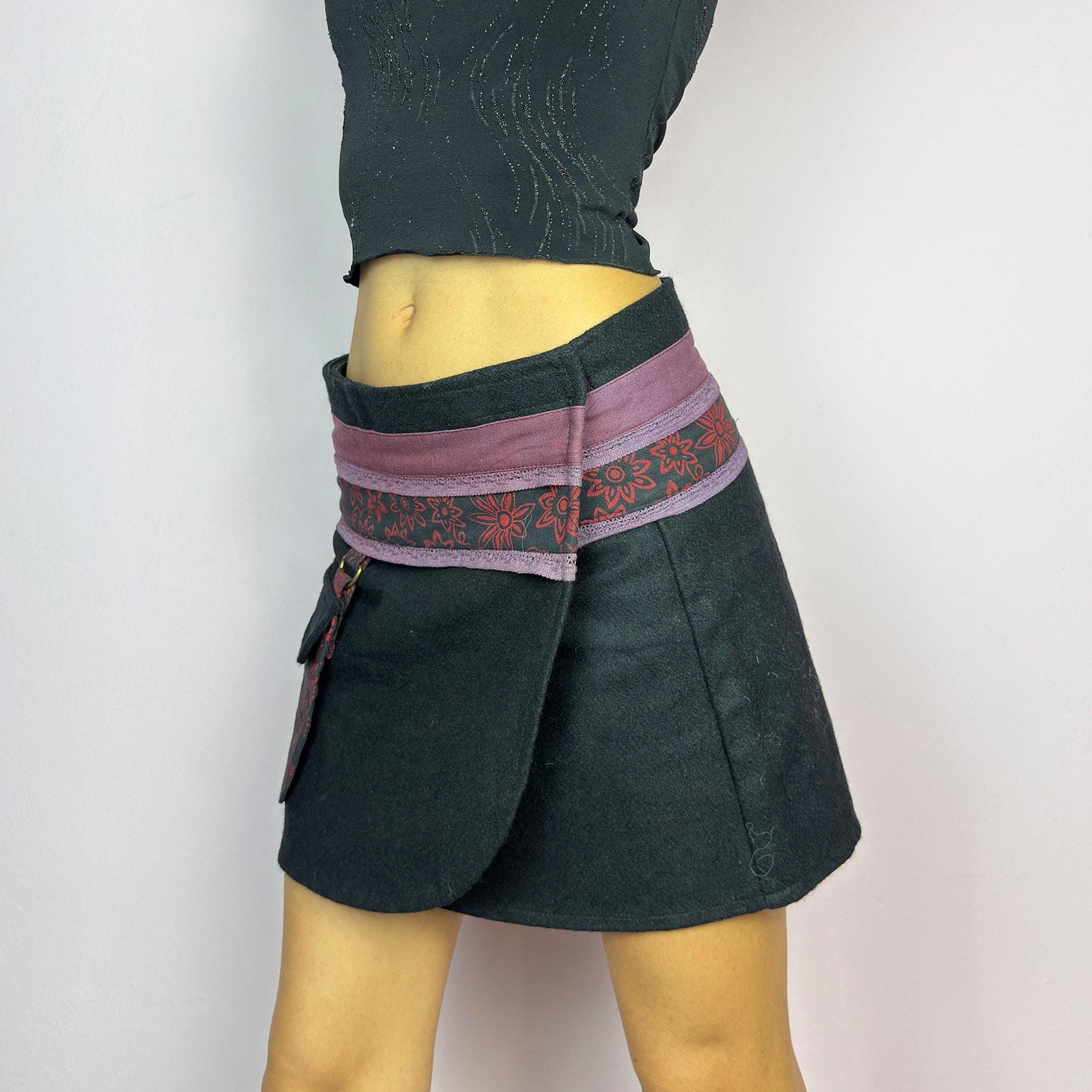 The Y2K Black Wrap Mini Skirt is a vintage 2000s piece featuring a unique wrap design with an adjustable Velcro waist for a customizable fit. Made from a thick fabric, the skirt has a contrasting waistband with floral details. adult world shop