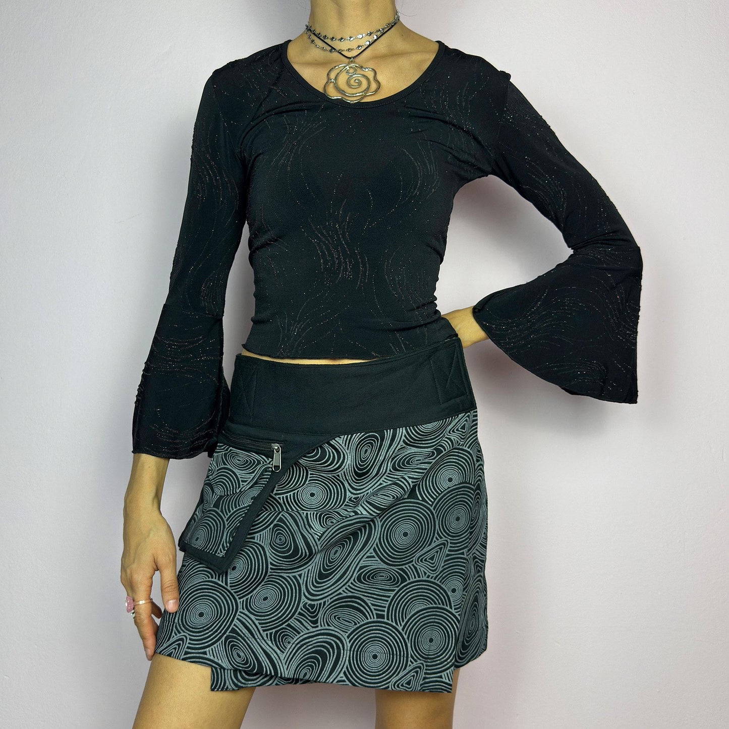 The Y2K Wrap Black Mini Skirt is a vintage 2000s piece designed with a Velcro wrap closure for an adjustable fit. Made from a thick fabric, it features a bold geometric swirl print and a utility pocket with a zipper closure. adult world shop