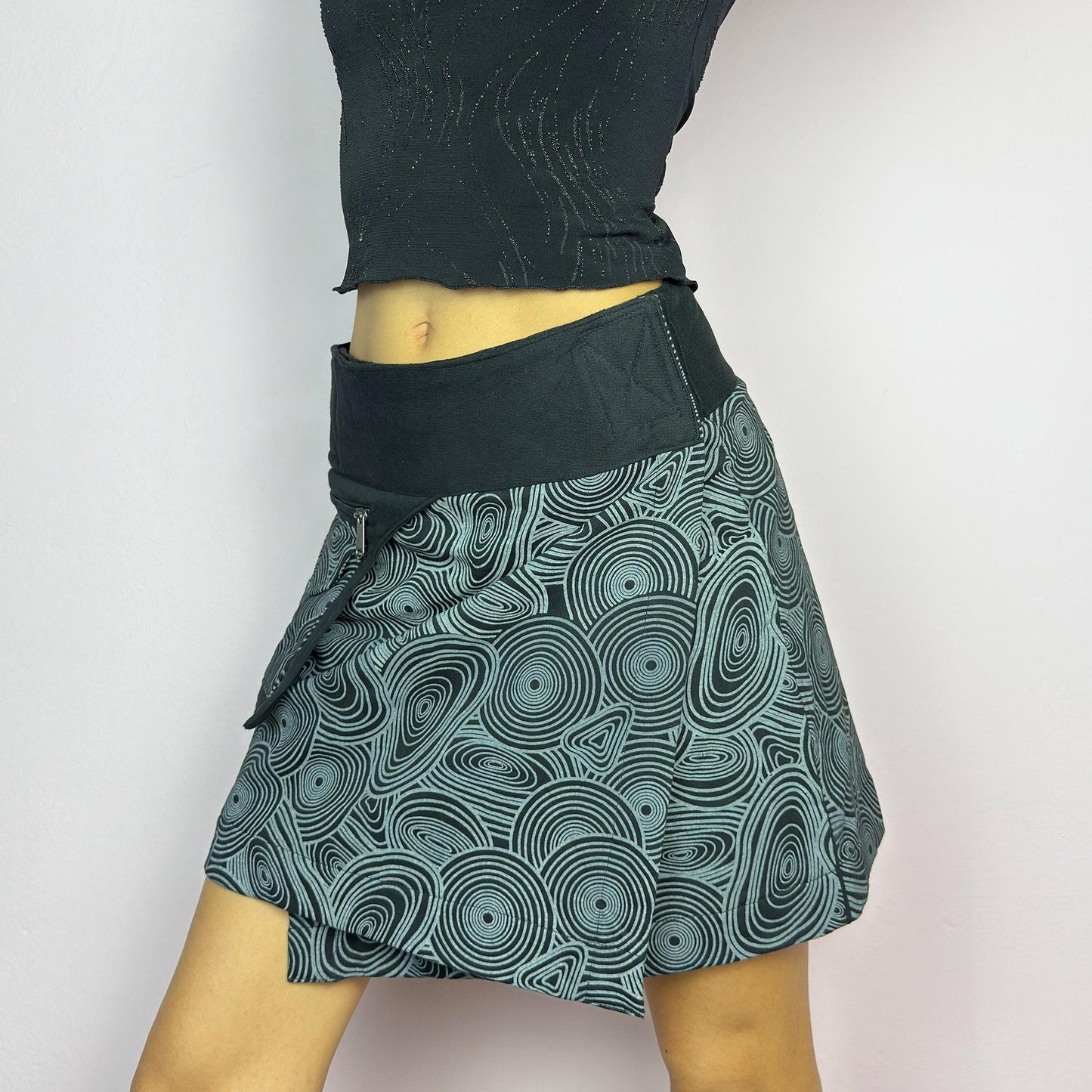 The Y2K Wrap Black Mini Skirt is a vintage 2000s piece designed with a Velcro wrap closure for an adjustable fit. Made from a thick fabric, it features a bold geometric swirl print and a utility pocket with a zipper closure. adult world shop