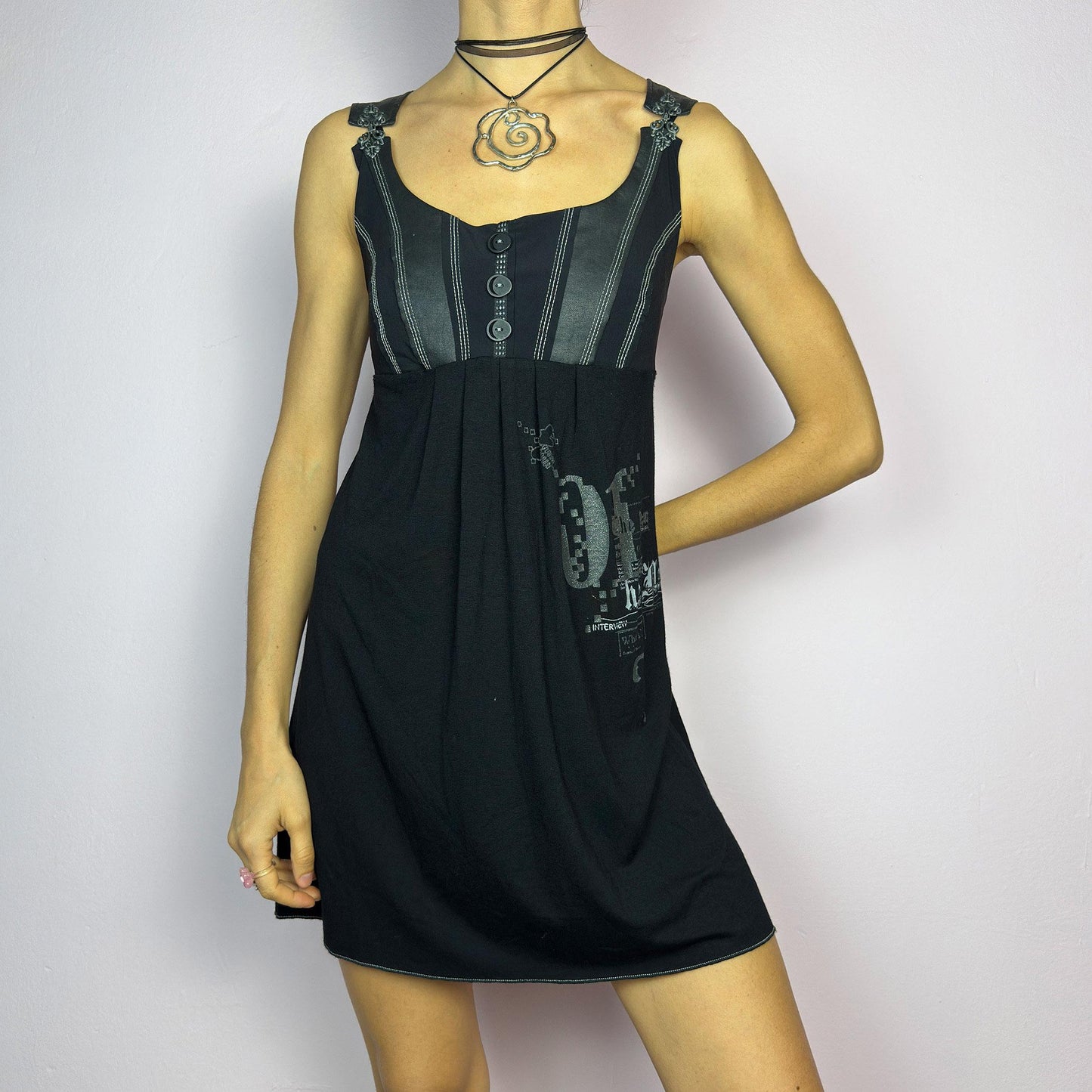 The Y2K Black Buckle Strap Mini Dress is a vintage 2000s sleeveless piece with a mix of edgy and casual aesthetics. It features decorative button details, contrast stitching, and graphic prints. adult world shop