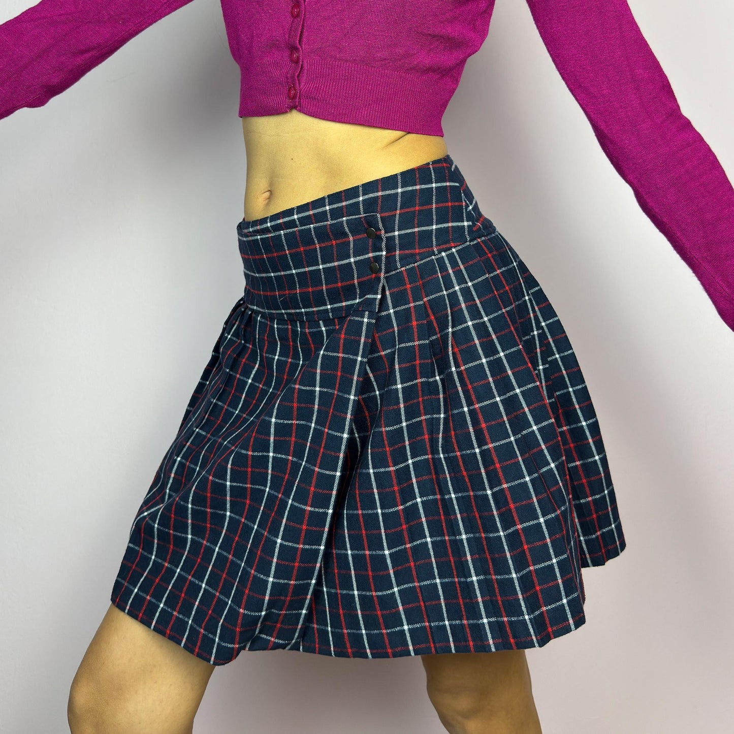 The Y2K Plaid Pleated Wrap Mini Skirt is a vintage 2000s piece featuring a wrap-style waistband with snap button details. Made from a plaid fabric in navy blue tones, it has structured pleats that create a classic silhouette. adult world shop