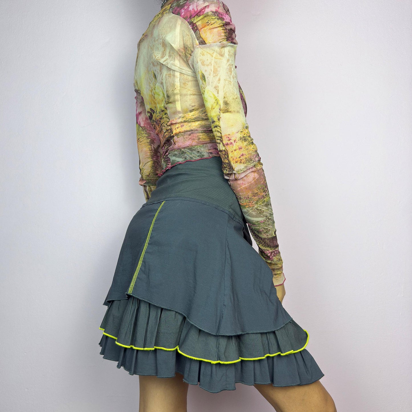 The Y2K Gray Ruffle Mini Skirt is a vintage 2000s statement piece featuring a tiered layered design with yellow contrast stitching details. Designed with a high-waist fit, it includes a side zipper closure. adult world shop