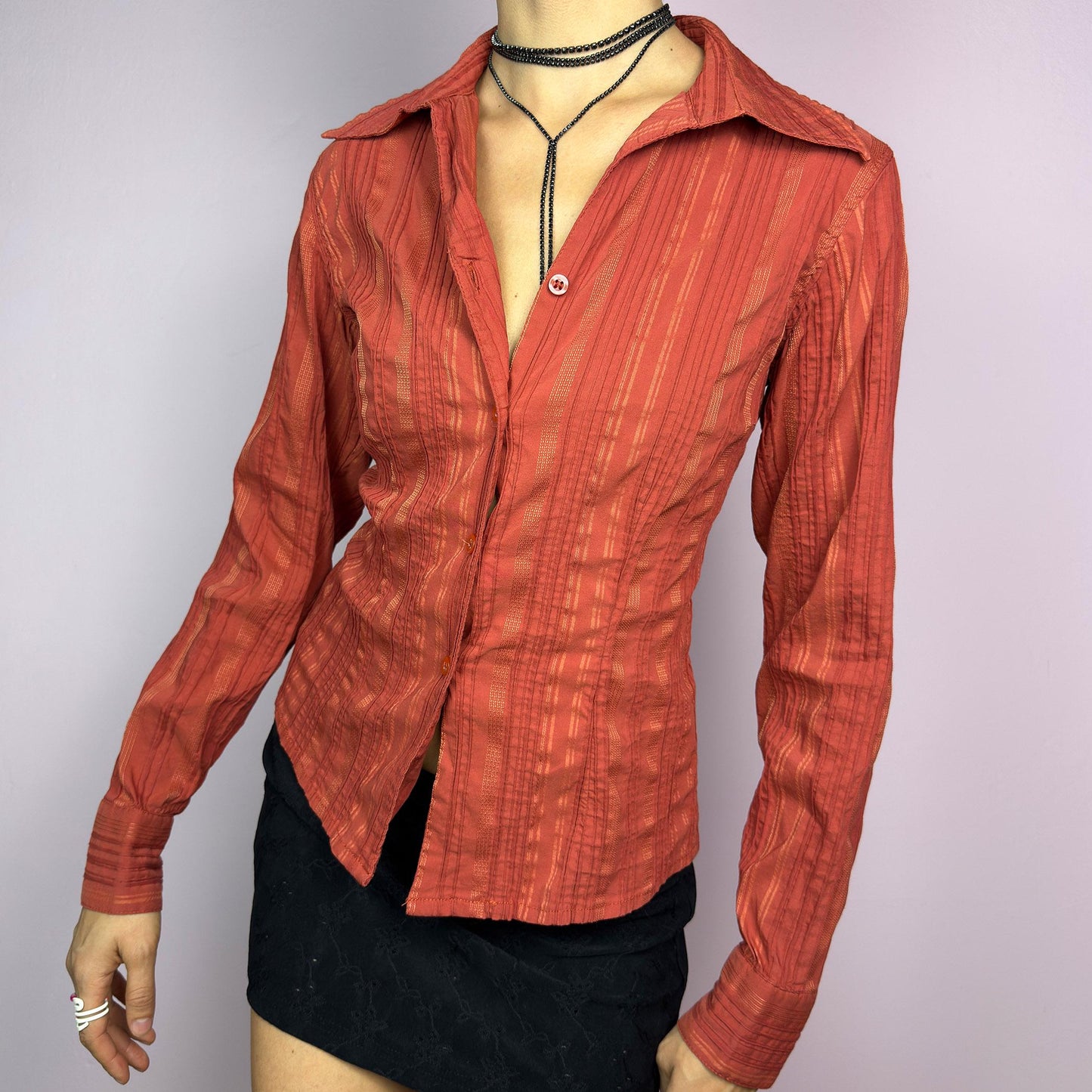 The Y2K Orange Striped Blouse is a vintage 2000s shirt featuring a warm rust-orange hue with a subtle striped pattern, a flattering tailored fit, and a hint of stretch. This long-sleeve button-up blouse is perfect for office wear. adult world shop