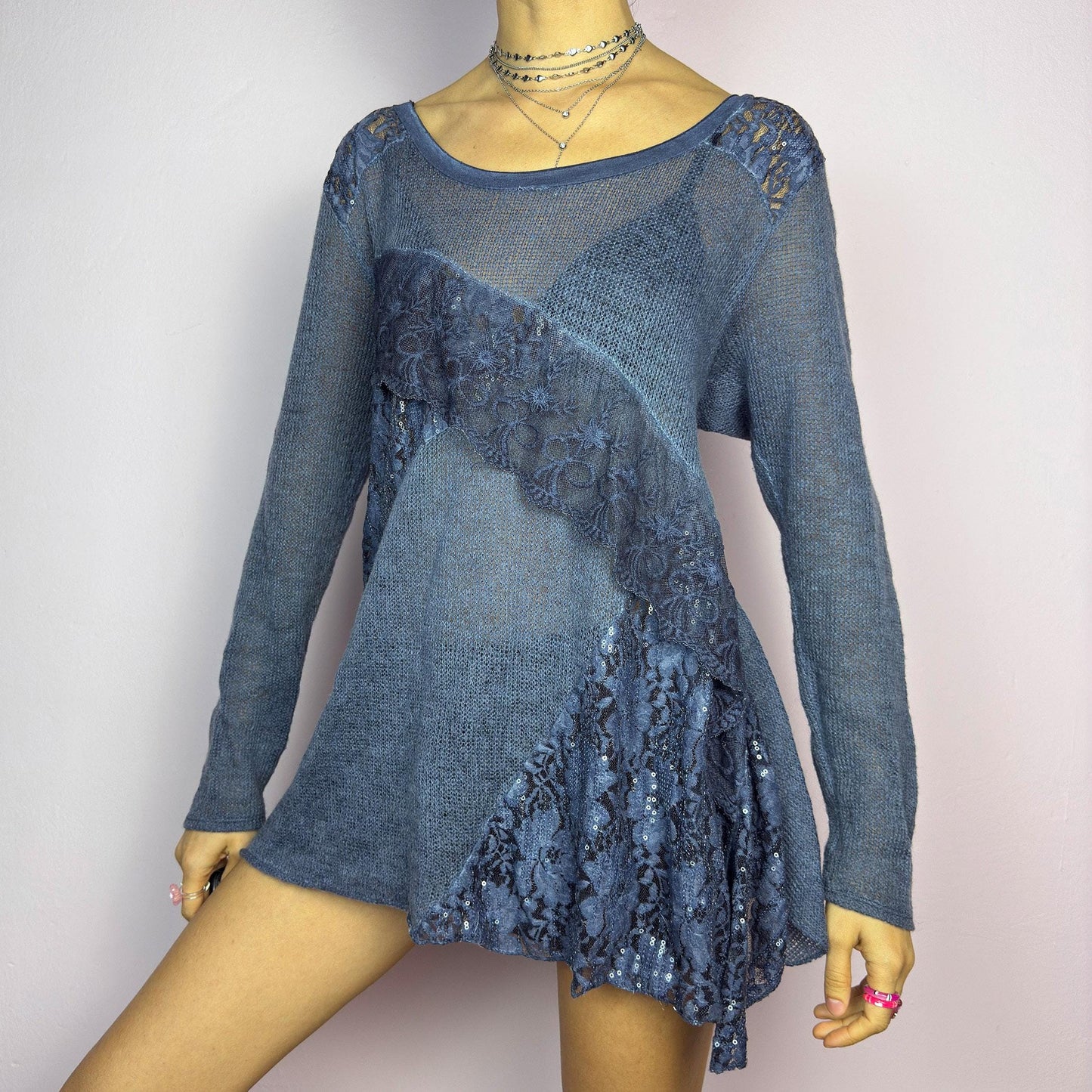 The Y2K Blue Lace Asymmetric Sweater is a vintage 2000s piece featuring a semi-sheer knit design with delicate lace and sequin paneling in an asymmetrical pattern. Designed with a scoop neckline, long sleeves, and a flowy silhouette, this sweater tunic adds a dreamy, romantic touch to any outfit. Perfect for layering in autumn and winter. adult world shop