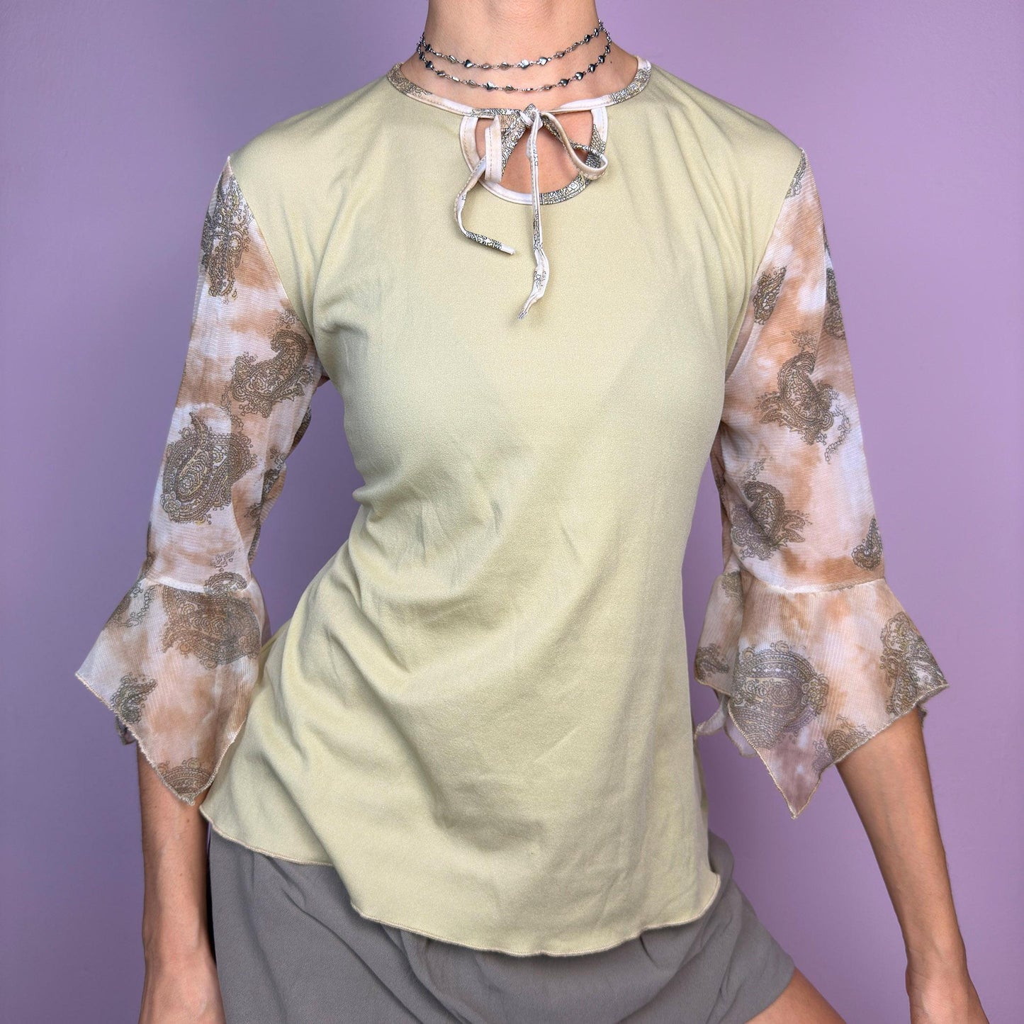 The Y2K Beige Bell Sleeve Tie Top is a vintage 2000s piece featuring a beige body with sheer paisley-printed bell sleeves in warm brown tones. Designed with a keyhole tie neckline, this dreamy top blends boho and romantic aesthetics. Perfect for casual, spring, and festival-inspired outfits. adult world shop