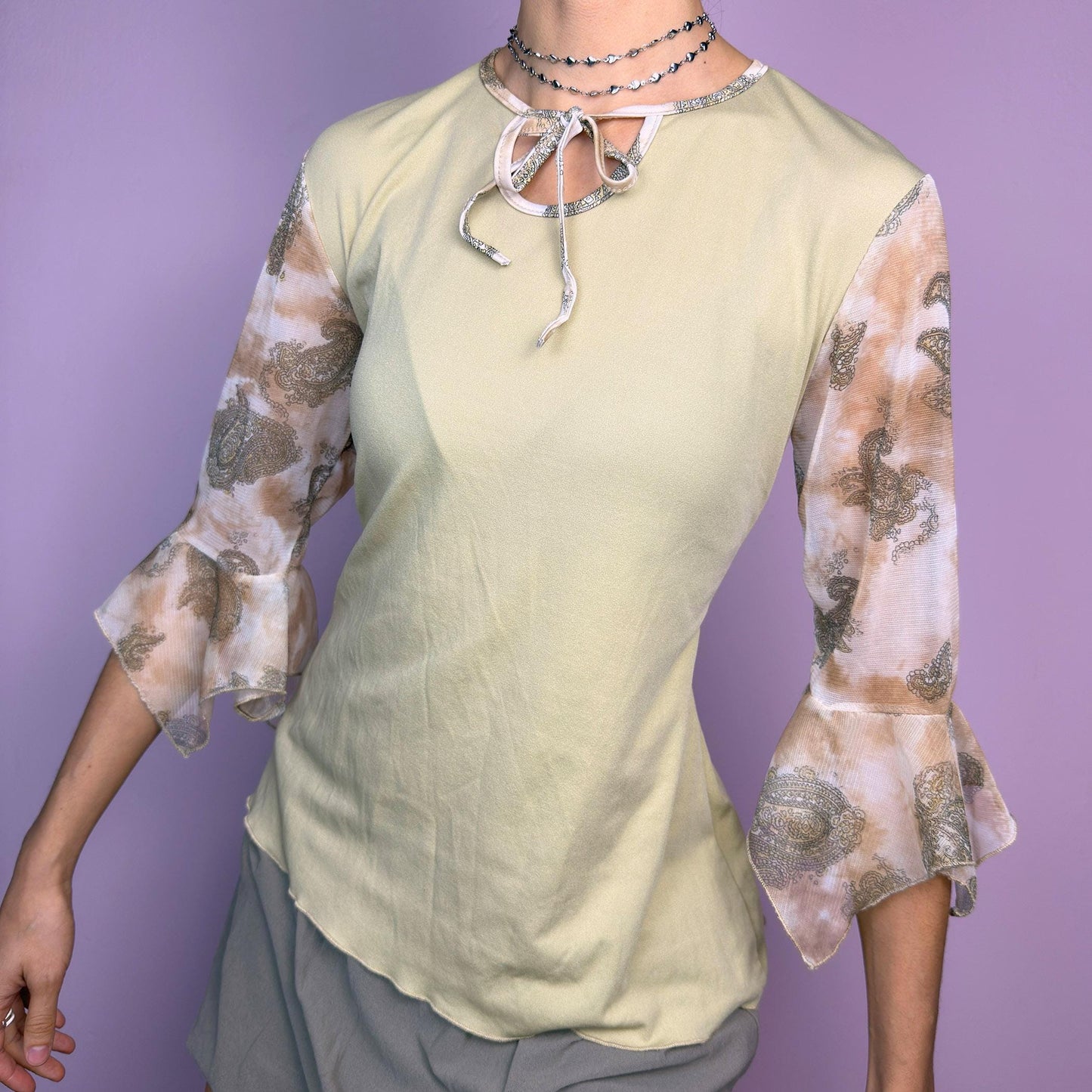 The Y2K Beige Bell Sleeve Tie Top is a vintage 2000s piece featuring a beige body with sheer paisley-printed bell sleeves in warm brown tones. Designed with a keyhole tie neckline, this dreamy top blends boho and romantic aesthetics. Perfect for casual, spring, and festival-inspired outfits. adult world shop