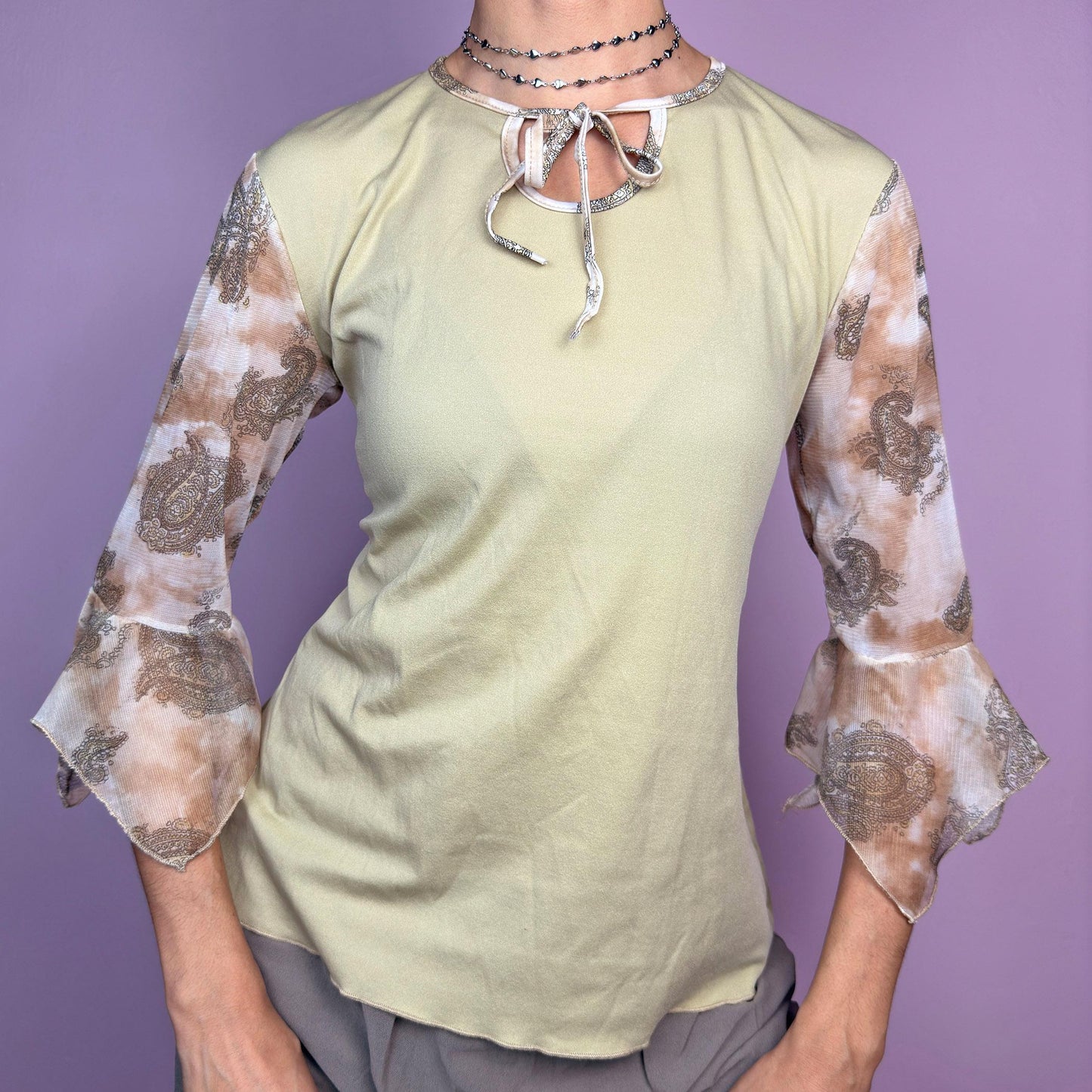 The Y2K Beige Bell Sleeve Tie Top is a vintage 2000s piece featuring a beige body with sheer paisley-printed bell sleeves in warm brown tones. Designed with a keyhole tie neckline, this dreamy top blends boho and romantic aesthetics. Perfect for casual, spring, and festival-inspired outfits. adult world shop
