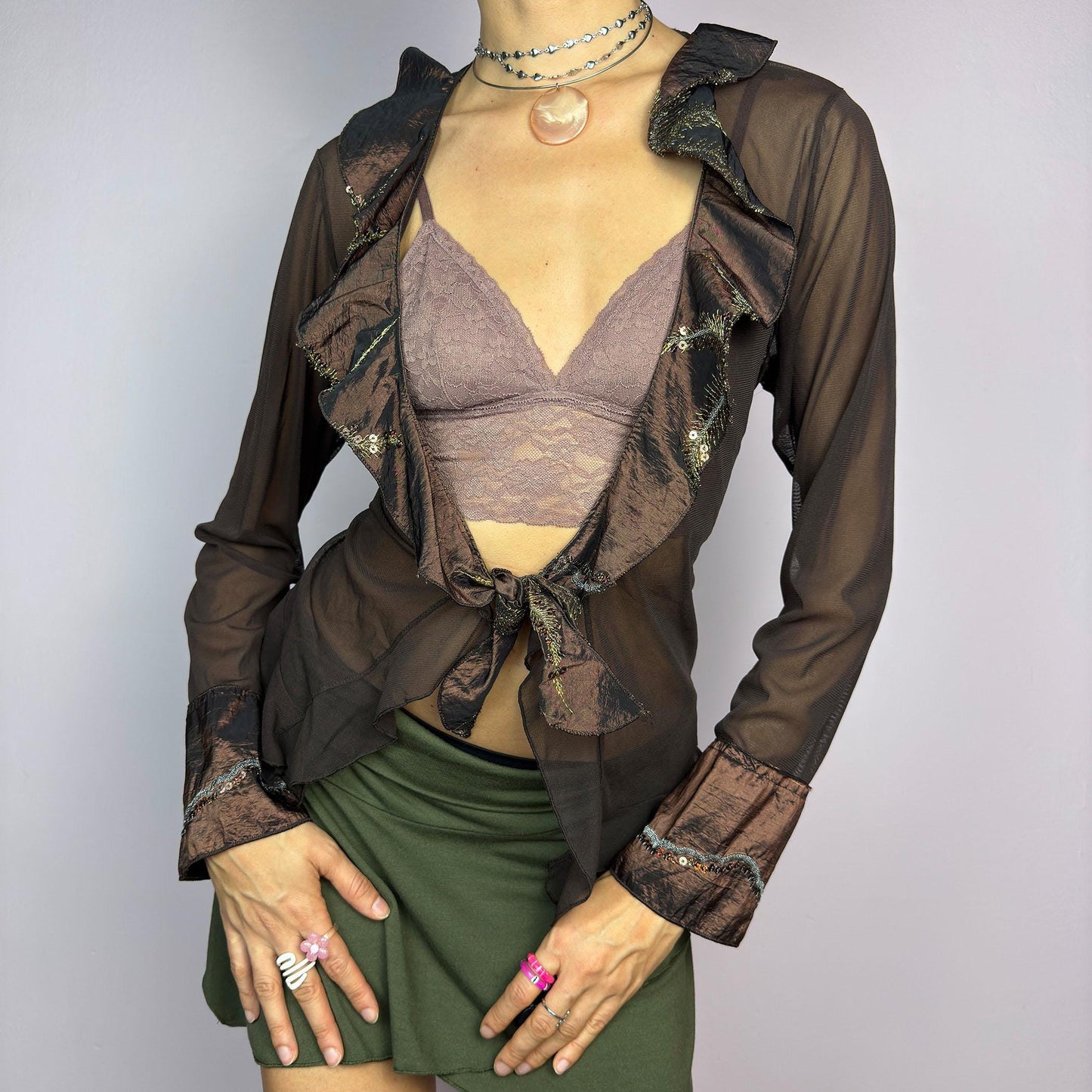 The Y2K Brown Mesh Tie Front Top is a vintage 2000s elegant and flirty piece. This semi-sheer mesh blouse features a tie-front closure and a cascade of ruffle detailing along the neckline and cuffs. adult world shop