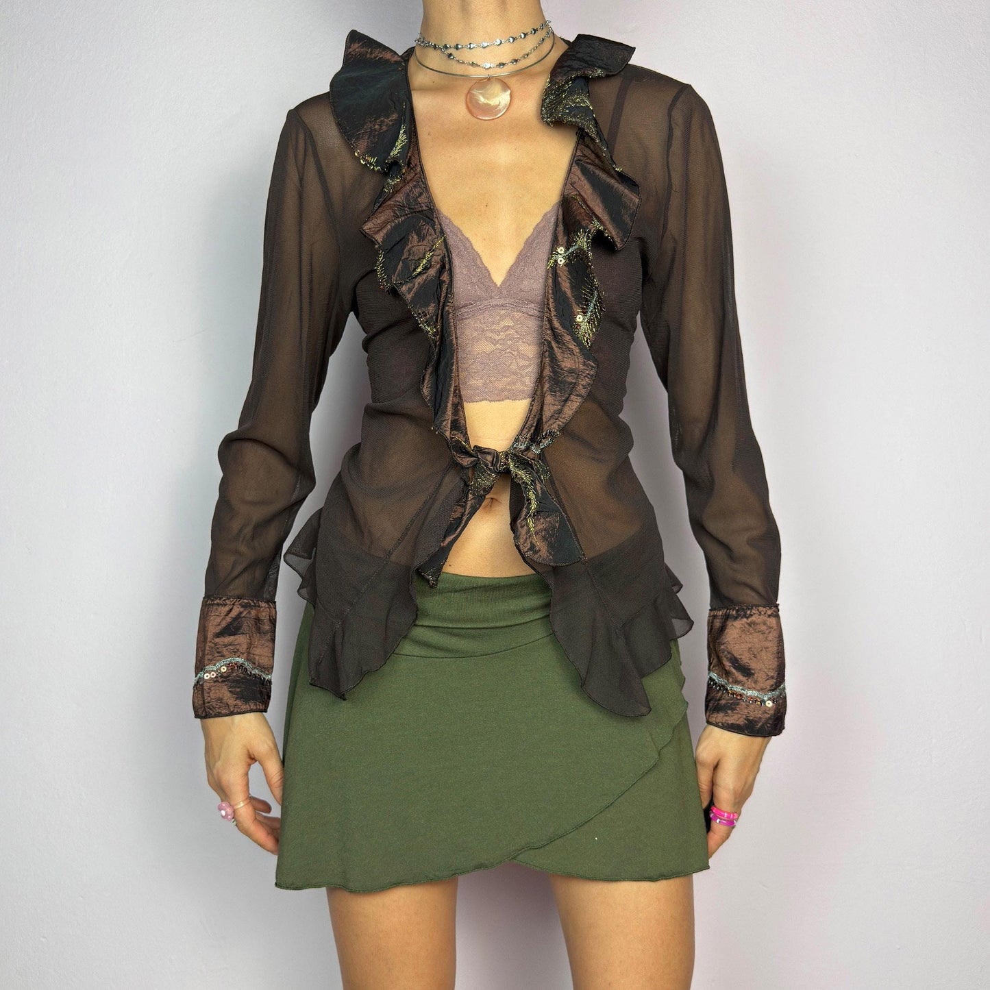 The Y2K Brown Mesh Tie Front Top is a vintage 2000s elegant and flirty piece. This semi-sheer mesh blouse features a tie-front closure and a cascade of ruffle detailing along the neckline and cuffs. adult world shop