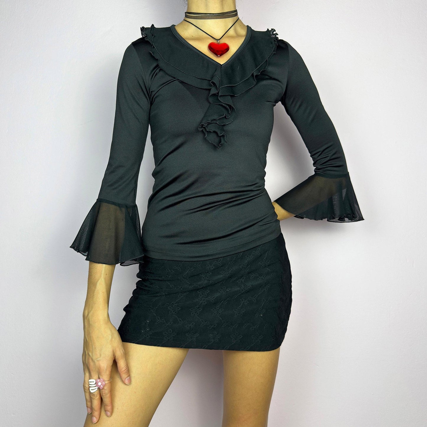 The Y2K Black Ruffle Bell Sleeve Top is a sleek and elegant vintage 2000s piece. Featuring a fitted silhouette with cascading ruffles along the neckline and a flattering V-neck design. The three-quarter bell sleeves with sheer mesh. adult world shop