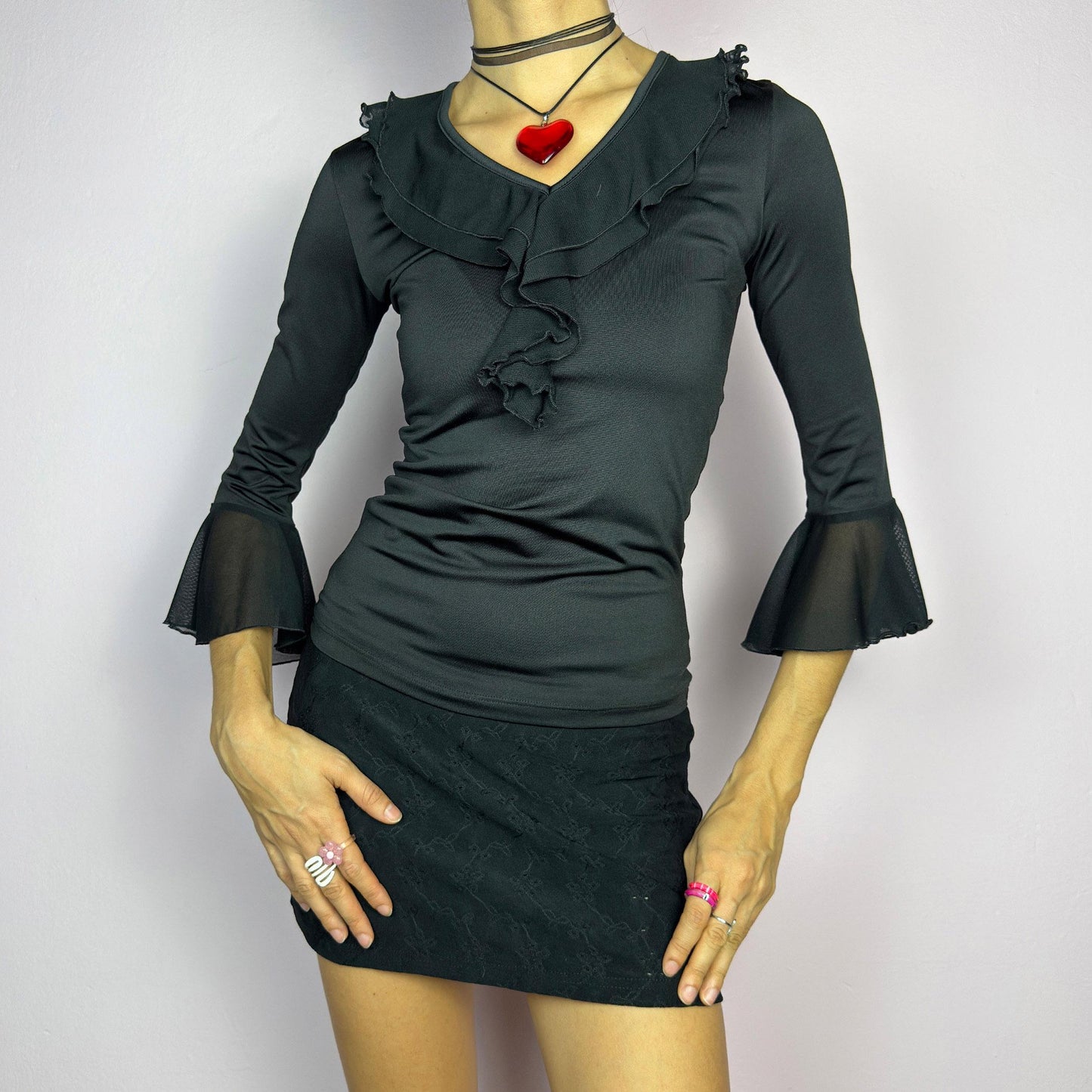 The Y2K Black Ruffle Bell Sleeve Top is a sleek and elegant vintage 2000s piece. Featuring a fitted silhouette with cascading ruffles along the neckline and a flattering V-neck design. The three-quarter bell sleeves with sheer mesh. adult world shop