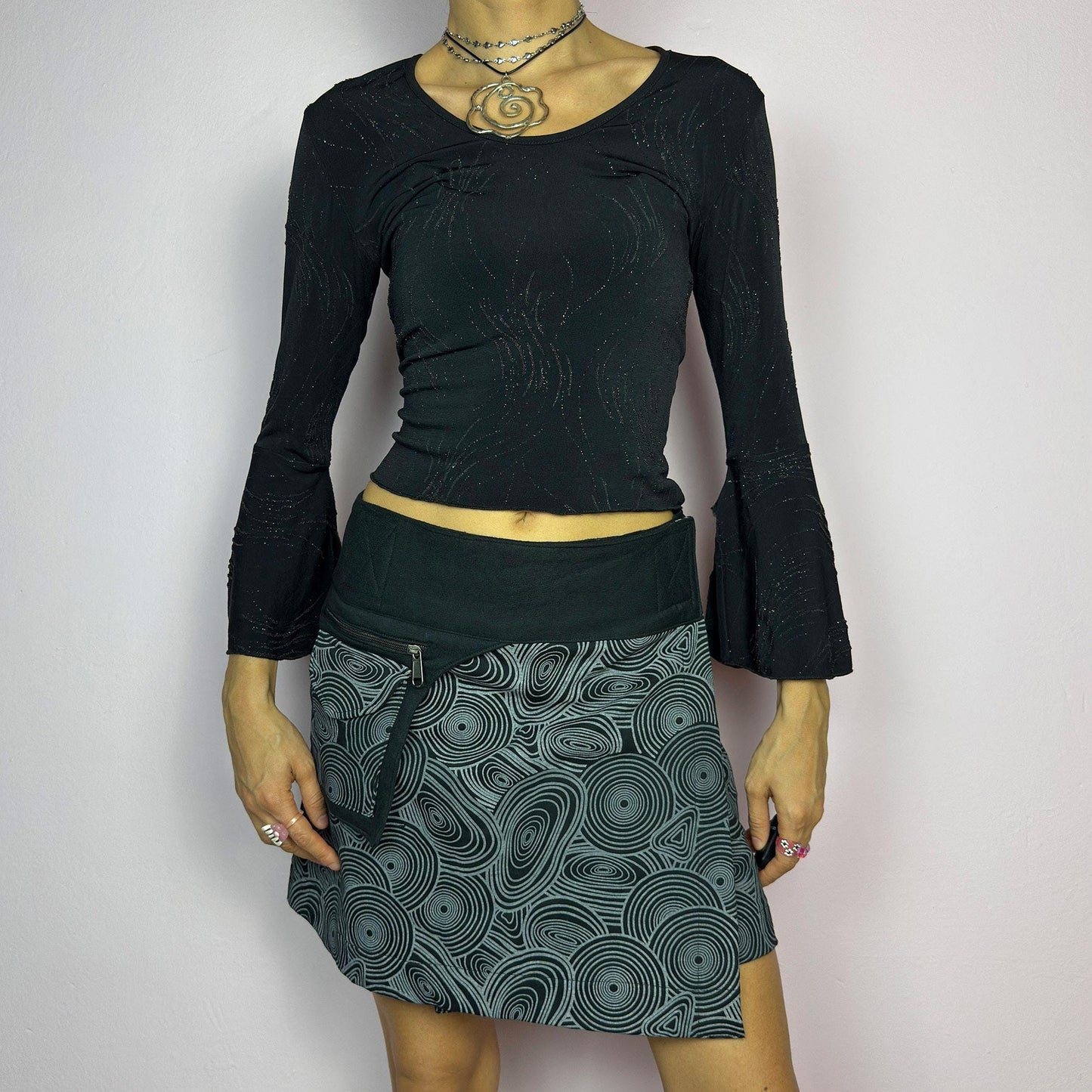 The Y2K Wrap Black Mini Skirt is a vintage 2000s piece designed with a Velcro wrap closure for an adjustable fit. Made from a thick fabric, it features a bold geometric swirl print and a utility pocket with a zipper closure. adult world shop
