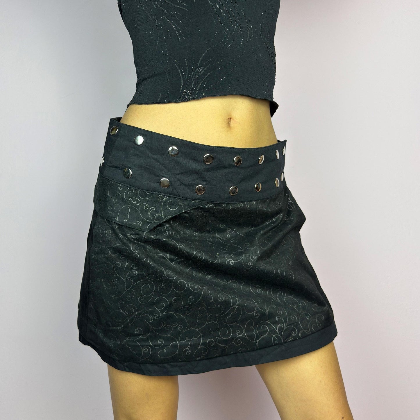 The Y2K Wrap Mini Black Skirt is a vintage 2000s piece featuring a snap-button closure on the waistband for an adjustable fit. Made from a thick fabric with a subtle floral pattern design, its wrap silhouette. adult world shop