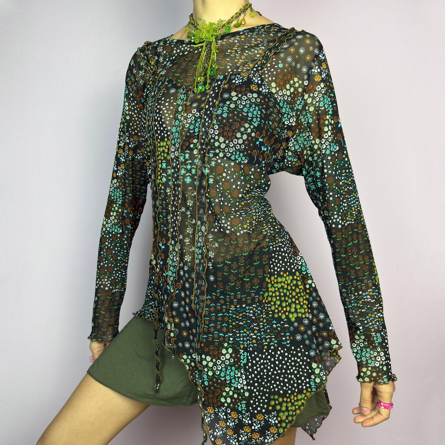 The Y2K Black Asymmetric Mesh Top is a vintage 2000s statement piece featuring semi-sheer mesh fabric with a floral and geometric print. Designed with an edgy asymmetric cut and delicate lettuce hem detailing, this top embodies a deconstructed aesthetic with a fairy grunge-inspired touch. The lightweight, stretchy fabric makes it perfect for layering, making it an ideal choice for summer avant-garde outfits. adult world shop