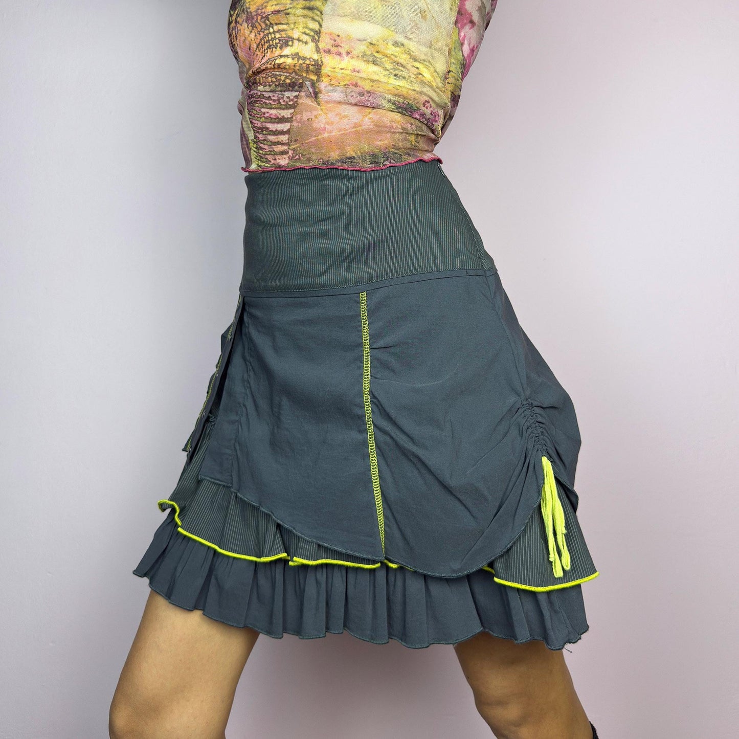 The Y2K Gray Ruffle Mini Skirt is a vintage 2000s statement piece featuring a tiered layered design with yellow contrast stitching details. Designed with a high-waist fit, it includes a side zipper closure. adult world shop